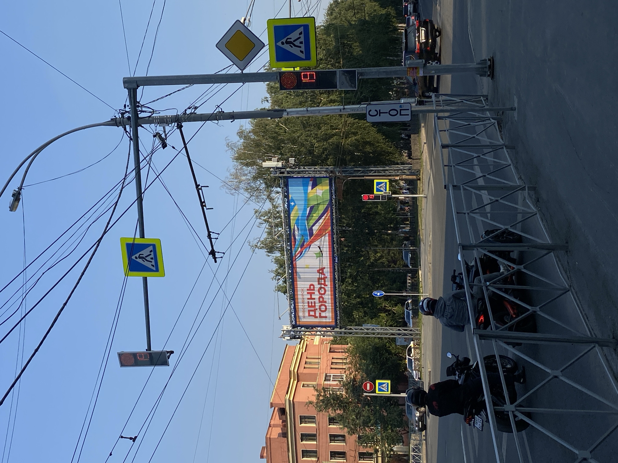 The image depicts an urban street scene in Novosibirsk, Russia, characterized by a clear blue sky and lush green trees. At the intersection, a large digital billboard displays a colorful graphic with Russian text, including "ДЕНЬ ГОРОДА" (City Day). Traffic lights and pedestrian crossing signs are visible, along with a prominent "STOP" sign. Two individuals on motorcycles are waiting at the intersection, one in a black jacket and the other in a gray hooded sweatshirt, both wearing helmets. Several cars are present on the road, indicating ongoing traffic. The architecture in the background includes a multi-story pink building with arched windows. The foreground features a metal fence. The overall impression is a bustling city intersection during daytime, likely commemorating a local event.