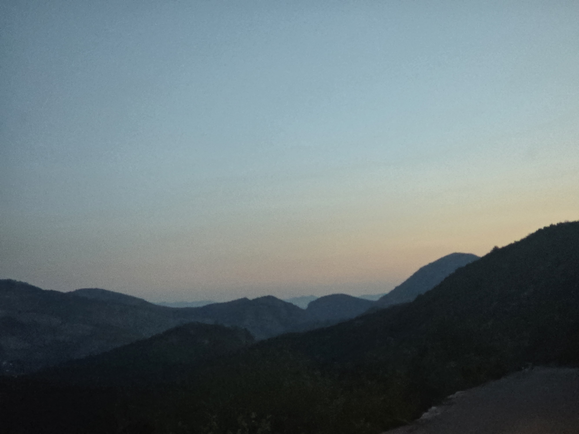 The image captures a panoramic view of mountainous terrain at dusk. The sky is a gradient of pale blue at the top, transitioning to a soft, peachy hue near the horizon, indicating the setting of the sun. Layered in the foreground and midground are dark, silhouetted mountain ranges, their contours softened by the dim light. The closest mountain range in the foreground is densely covered with dark vegetation. Further back, additional mountain ranges are visible, becoming progressively fainter and bluer with distance, suggesting atmospheric perspective. The overall scene is serene and natural, with no visible man-made structures or human presence. The location is identified as the vicinity of Klopot, Montenegro.