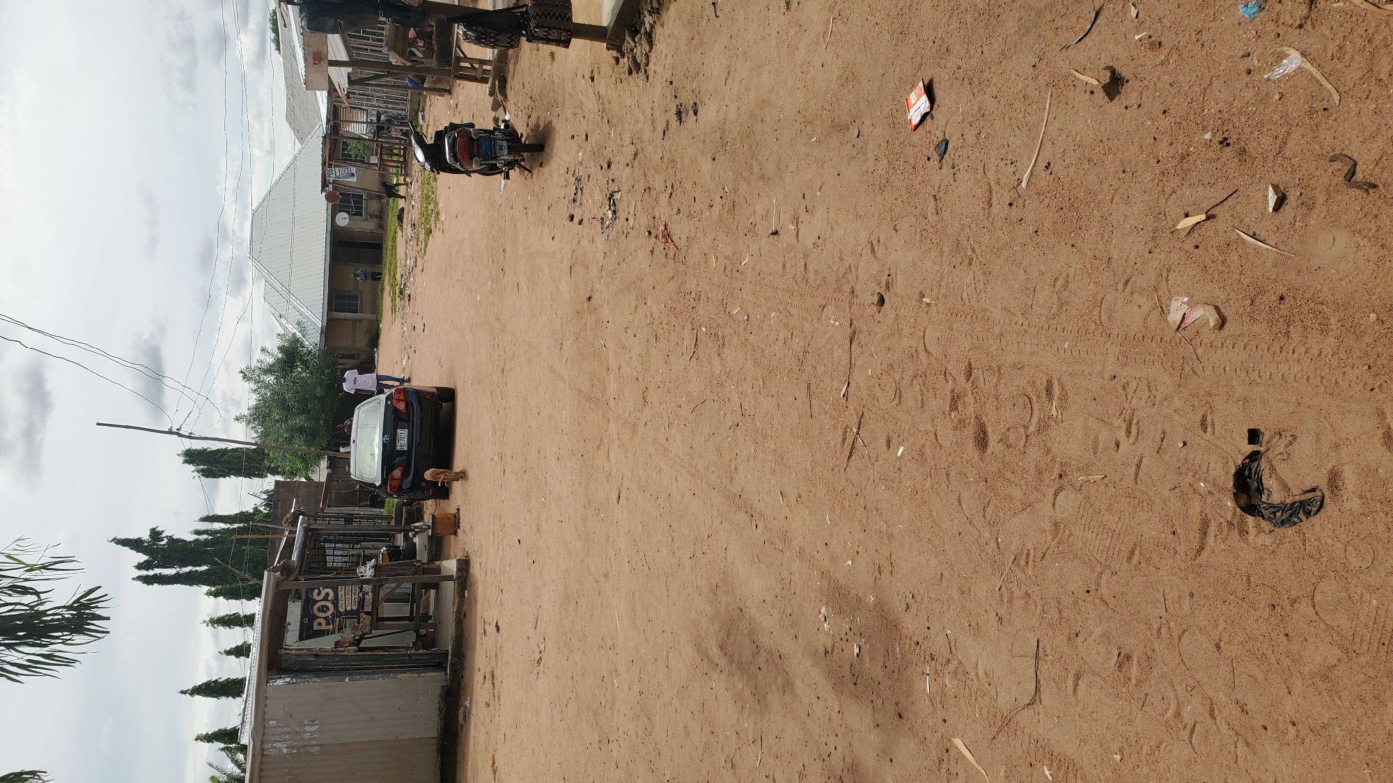 A vertical, eye-level, outdoor, medium shot captures a dirt road in Jalingo, Nigeria, under an overcast sky. The road is primarily composed of light brown sand with tire tracks and scattered debris, including leaves, twigs, and pieces of trash. On the left, a corrugated metal shack with an open front displays a sign that reads "POS." A black sedan with a license plate reading "HBC 781J" is parked in front of the shack. A small, light-brown dog is standing near the rear of the car. Further down the road, a person in a white t-shirt and dark shorts walks away from the camera. On the right side of the road, a black motorcycle is parked. In the background, several buildings with metal roofs are visible, along with tall, thin trees and overhead power lines. Another person, wearing a blue shirt, stands near one of the background buildings. The scene conveys a sense of everyday life on a dusty road.