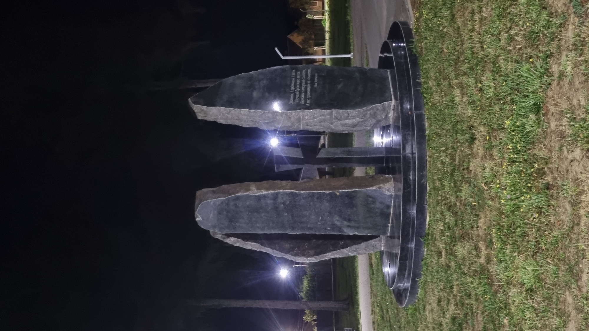 The image shows a monument at night, illuminated by streetlights. The monument consists of three large, dark, abstract stone slabs arranged in a semi-circle, with a dark, polished circular base. In the center of the semi-circle, a dark, polished cross stands vertically. The stone slabs have rough, natural edges and smooth inner surfaces. Inscriptions are visible on the rightmost slab, in Cyrillic script. The monument is situated on a grassy area with some yellow wildflowers. In the background, trees and a building are visible, along with streetlights. The monument is identified as the "Monument to the Victims of Fascism" located in Mukachevo, Ukraine.