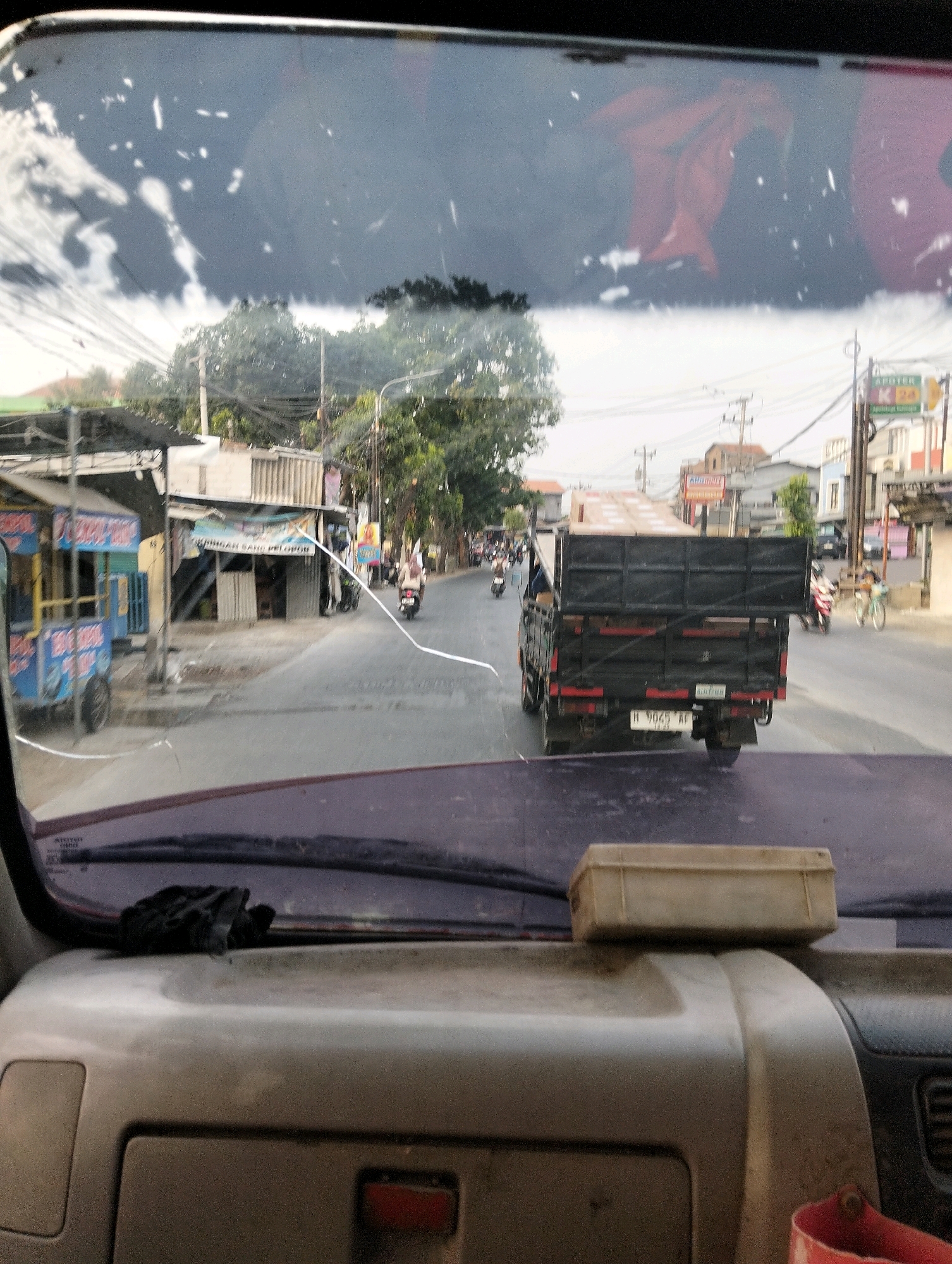 The image is taken from inside a vehicle, looking through the windshield. The windshield is cracked and smudged, with reflections of a reddish fabric or object visible.
Outside, a two-lane road runs through a commercial area. On the left side of the road, there are several small shops or stalls, one of which is blue and yellow with "BOBO" written on it, and another with a sign that says "SANG PELOPOR". Further down the road, there are trees and buildings, including what appears to be an "APOTEK K 24" sign. The sky is overcast.
Several vehicles are on the road. In the middle of the frame, a black flatbed truck with a red license plate and the registration number "H 9065 AF" is driving away from the camera. There are wooden planks loaded onto the truck bed. To the left of the truck, several motorcycles are visible, some with riders. To the right of the truck, a person on a bicycle is also visible.
In the foreground, inside the vehicle, the dashboard is visible. A black glove is placed on the dashboard, and a beige rectangular box is resting on a raised section of the dashboard. A red object, possibly a cup or container, is visible in the lower right corner.
The overall impression is a view from a vehicle driving through a busy, somewhat rundown commercial area in Semarang, Indonesia, during daylight hours.