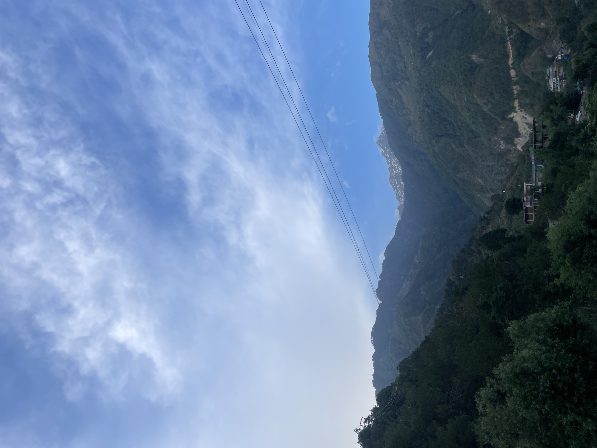 The image shows a mountain range in Dharamshala, India under a blue sky with scattered clouds. Power lines stretch across the sky towards the mountains. Trees and buildings are visible on the lower slopes of the mountains.