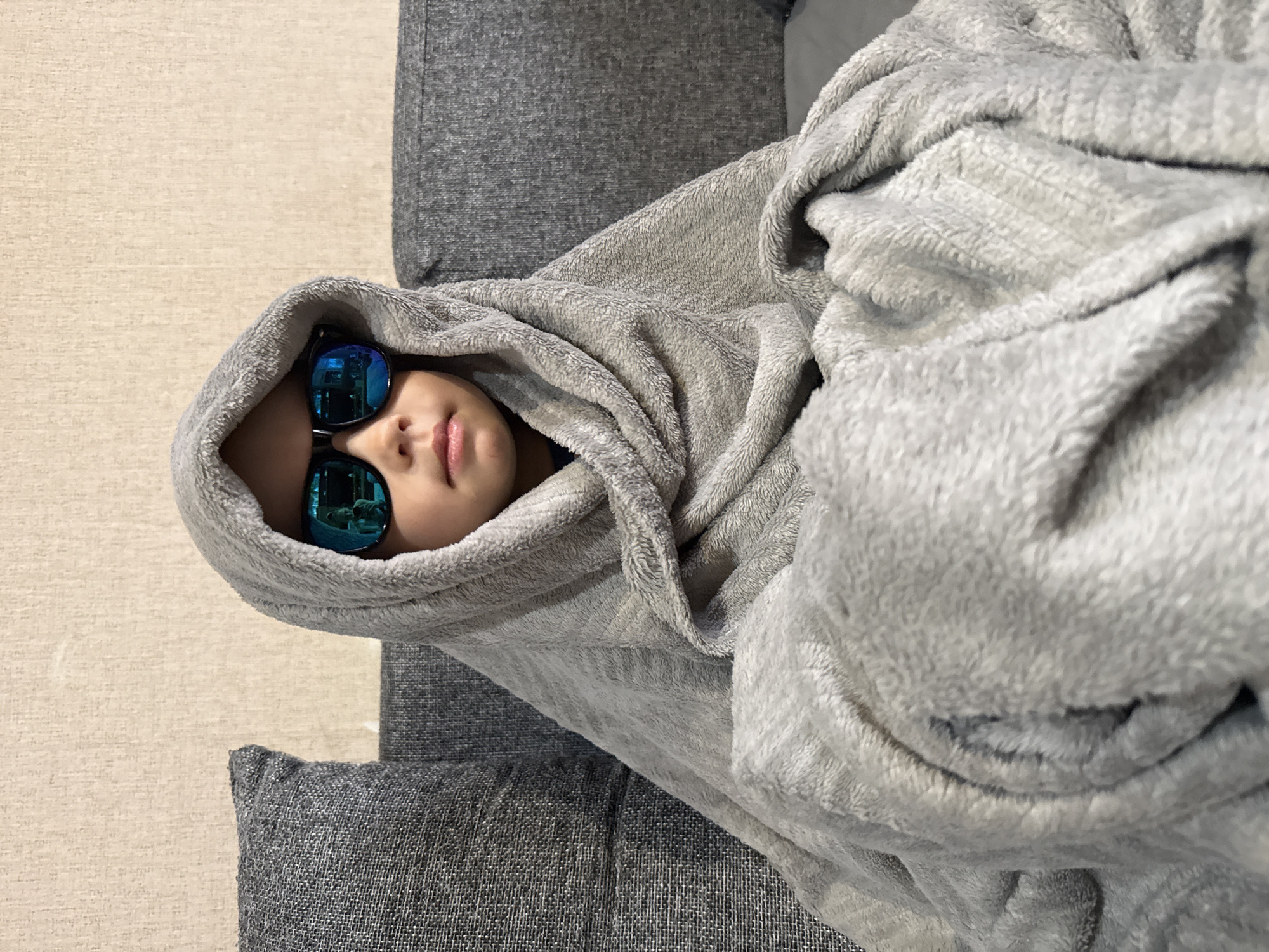 A child lies on a gray patterned couch, bundled in a soft, light gray blanket. The blanket is draped over their head, forming a hood. They are wearing sunglasses with reflective blue lenses. The background consists of a light tan carpet and a portion of the gray couch. It is not possible to confirm or deny the image's location in Rusanovo, Russia with the information present in the image.