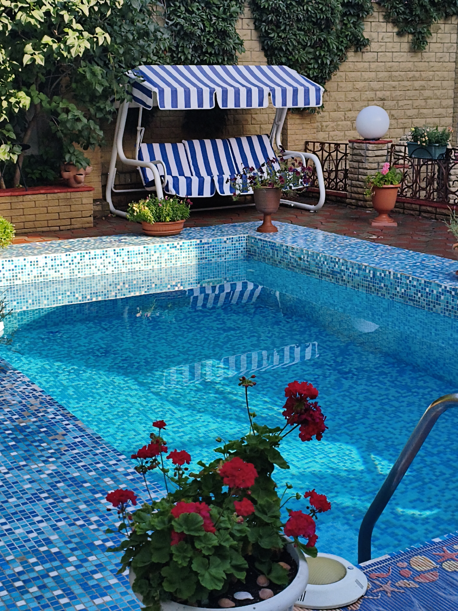 The image shows a small, tiled swimming pool with bright blue water. A swing set with a blue and white striped canopy and cushions is situated next to the pool. Several potted plants with red and yellow flowers are placed around the pool area. In the background, there is a brick wall covered in ivy and a decorative sphere on a stone pedestal. The scene suggests a private backyard or patio setting. The city of Makiivka, Ukraine, is not discernible from the image itself.