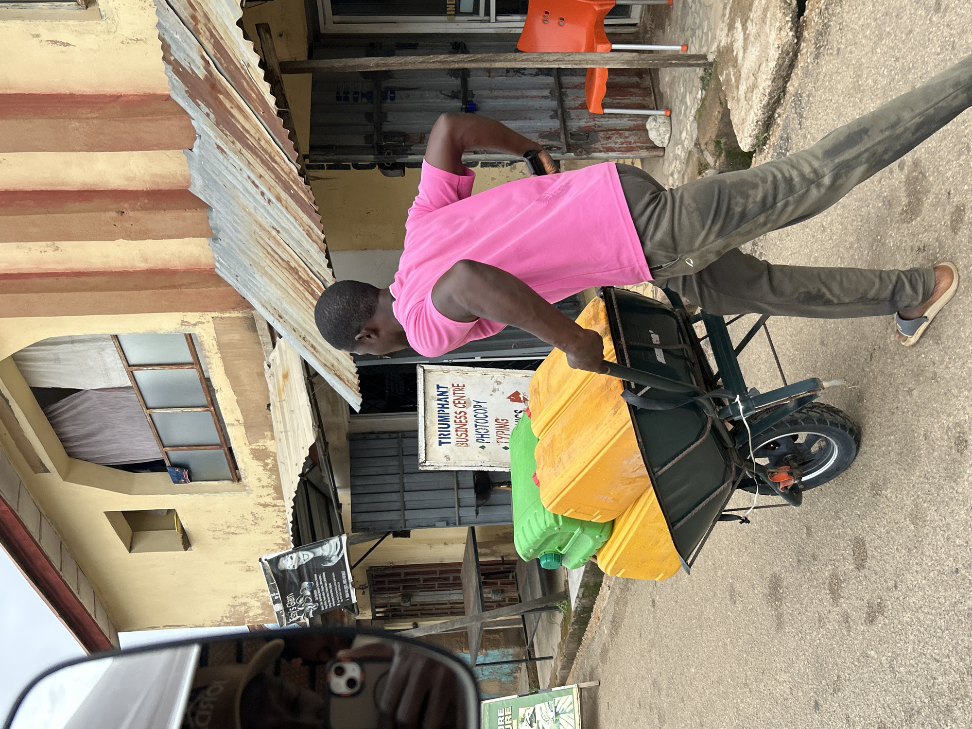 A man is shown pushing a wheelbarrow loaded with yellow and green jerrycans on a paved road. The location is Jos, Nigeria, as indicated by the "Triumphant Business Centre" sign visible in the background. The man is wearing a pink shirt and dark pants, and light-colored sandals. He is in motion, pushing the wheelbarrow away from the camera's perspective. The background includes buildings with weathered facades and corrugated metal roofing. A glimpse of a side-view mirror, capturing a smartphone, is visible at the bottom left of the frame.