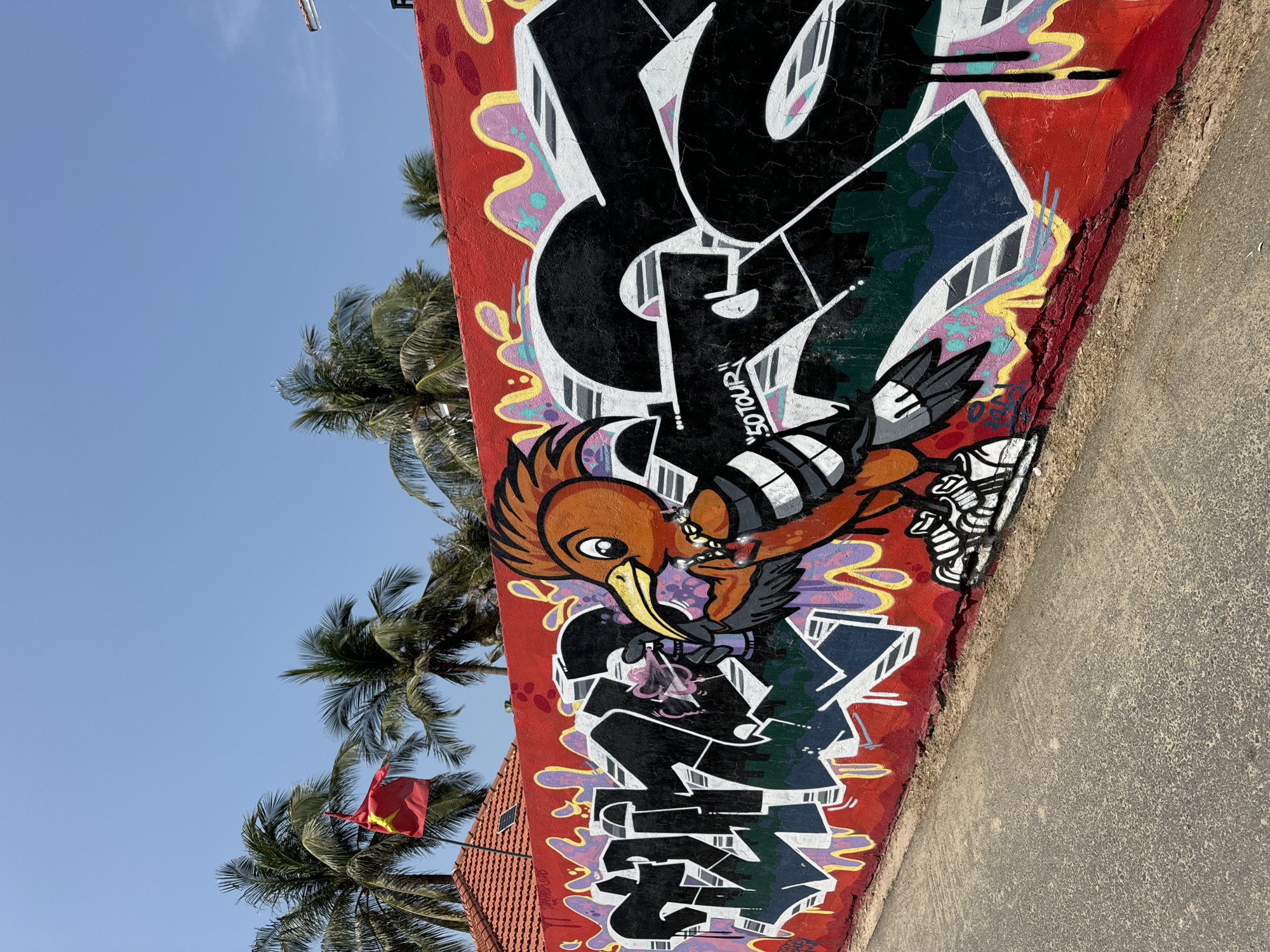 A street-level view depicts a segment of a concrete wall adorned with graffiti. The wall is painted red and covered with colorful graffiti art. An illustration of an orange bird, which is the main feature of the graffiti, is placed near the middle. The bird is carrying a beaded necklace with its beak. Palm trees are visible in the background against a clear blue sky. In the lower left corner, a Vietnamese flag is planted. The scene appears to be located in Mũi Né, Vietnam, indicated by the flag and the tropical landscape. Next to the bottom right of the wall lies a concrete paved road.