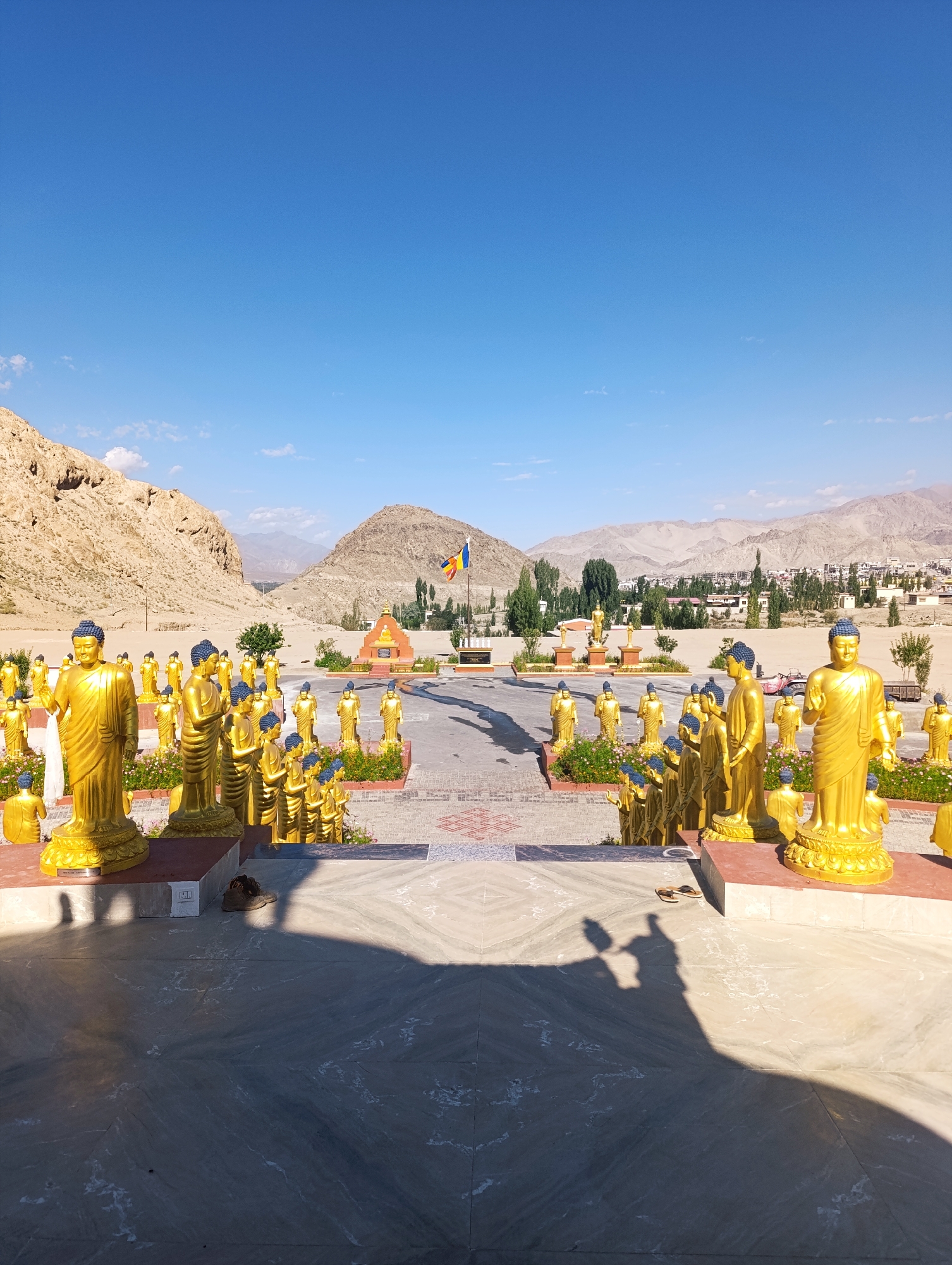 A wide, low-angle outdoor shot captures a courtyard filled with numerous golden statues of the Buddha, arranged in rows. The statues vary in size, with some standing and others appearing to be seated. They are golden, with blue caps and robes. In the background, a Buddhist temple structure with an orange roof and a flag on a pole are visible. Beyond the temple, a cityscape with buildings and trees is nestled between rugged, tan-colored mountains. The sky above is a clear, bright blue. In the foreground, a marbled courtyard floor is visible, with shadows cast across it. A pair of brown sandals rests on the floor near the right side of the frame. The scene appears to be the Maitreya Buddha statue at the Sankar Gompa monastery in Leh, India, given the context of golden Buddha statues and the mountainous terrain characteristic of Leh.