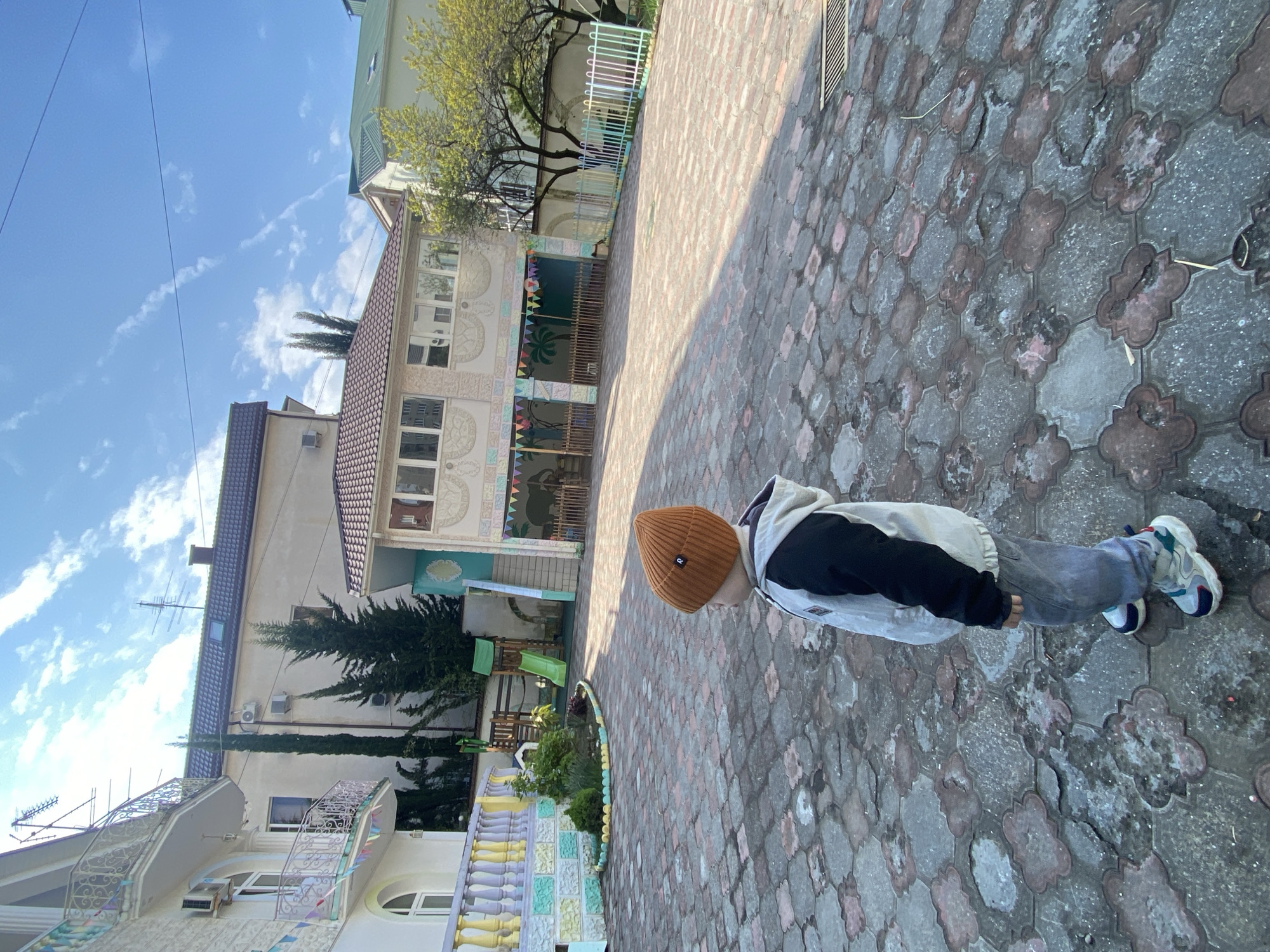 A full shot captures a person on a paved area, potentially in Sochi, Russia. The individual, clad in a brown beanie, a two-tone jacket (light gray and black), gray pants, and multi-colored sneakers, stands on cobblestone or interlocking brickwork.
The background reveals buildings of varying heights and architectural styles. One building features multiple stories with a brown-tiled roof, interspersed windows, and ornate decorative elements. Adjacent to it, another building displays a white facade with decorative railings and arches.
Further in the background, trees, including one with pale green foliage, are visible. The sky is a clear blue with scattered, wispy clouds.