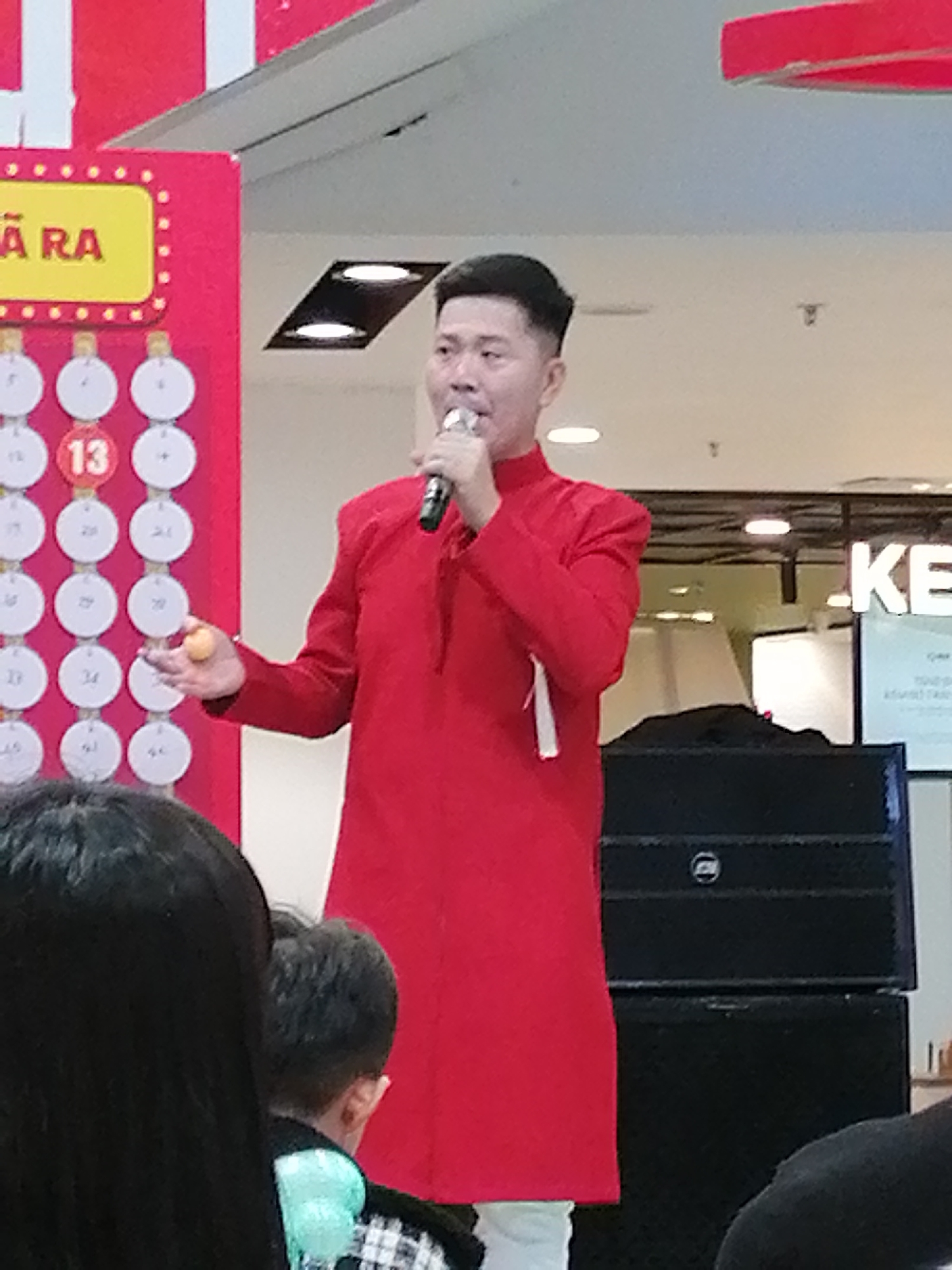 The image depicts a man in a red traditional Vietnamese outfit, likely an Áo dài, holding a microphone and a small orange ball. He appears to be addressing an audience, possibly during a promotional event or a lottery draw, given the presence of a large red board with numbered circles on the left side of the image. The board has a yellow sign with Vietnamese text, "RA" visible. The man's expression is animated, suggesting he is engaged in an energetic presentation.
Behind him, there are large black speakers, indicating a performance or an announcement is taking place. In the background, the word "KE" is visible in large white letters on a sign, possibly part of a store or business name. The setting appears to be indoors, likely a shopping mall or a public venue, with fluorescent lighting overhead.
The audience is partially visible in the foreground, with the back of a person's head and shoulders in dark hair, and another person with a black and white checkered shirt and a light blue scarf. The overall mood seems festive and lively, consistent with a public event in Da Lat, Vietnam. The time of day is not explicitly indicated, but the indoor lighting suggests it could be any time. The presence of a microphone and speakers points to an event involving speaking and possibly music or announcements. The small orange ball held by the man might be part of a game or a raffle.