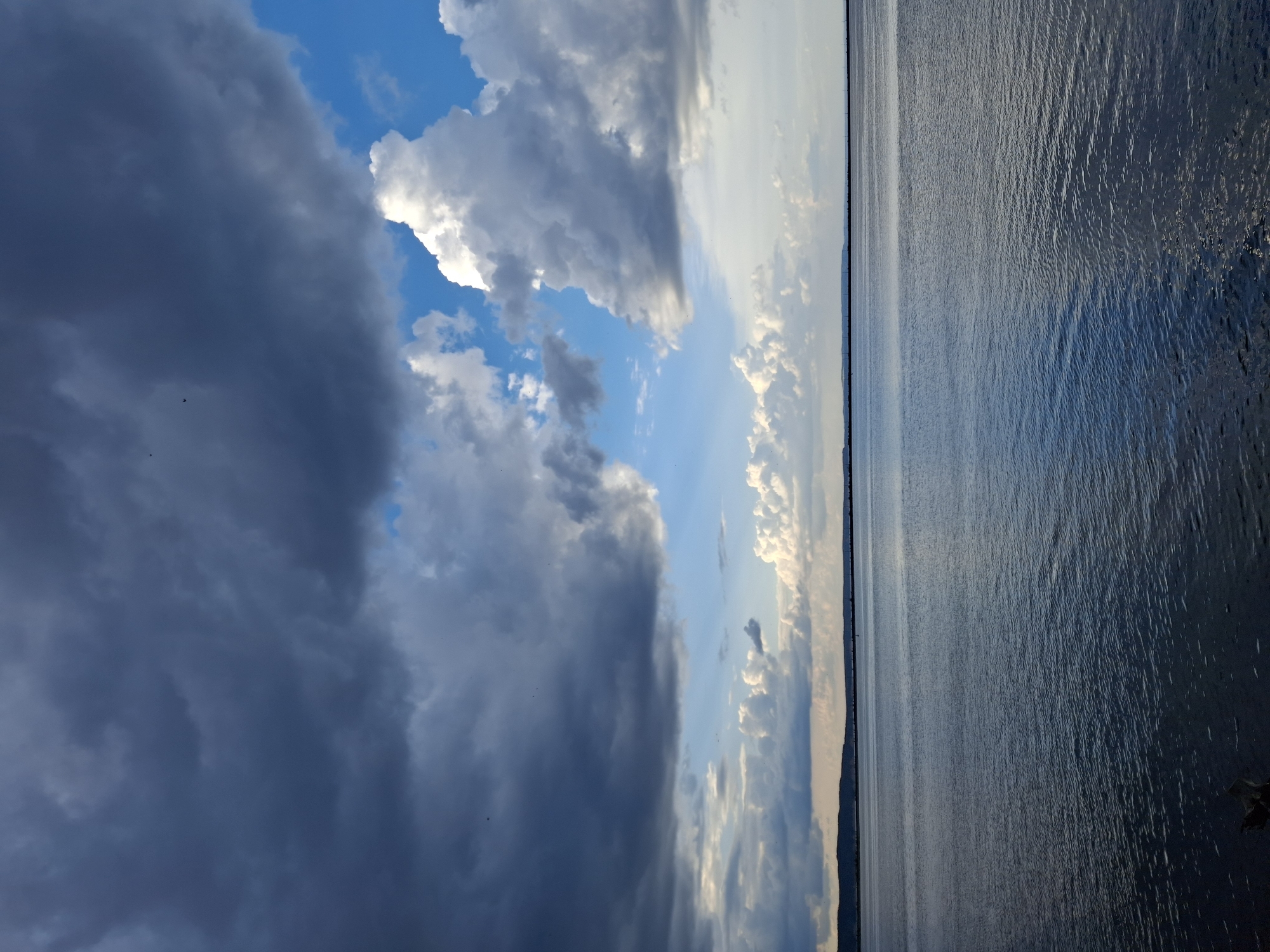 The image depicts a wide, calm body of water, likely a river or reservoir, with a distant, low-lying shoreline. The sky above is dramatic, filled with a mix of dark, heavy storm clouds and bright, illuminated cumulus clouds against patches of vivid blue sky. The light suggests either the approach or departure of a storm. There are no people or visible structures on the water or the shoreline. The scene is natural and unpopulated. This view is characteristic of the Kama River or its reservoir in the city of Chaikovsky, Russia, known for its extensive water expanse and often striking cloud formations.