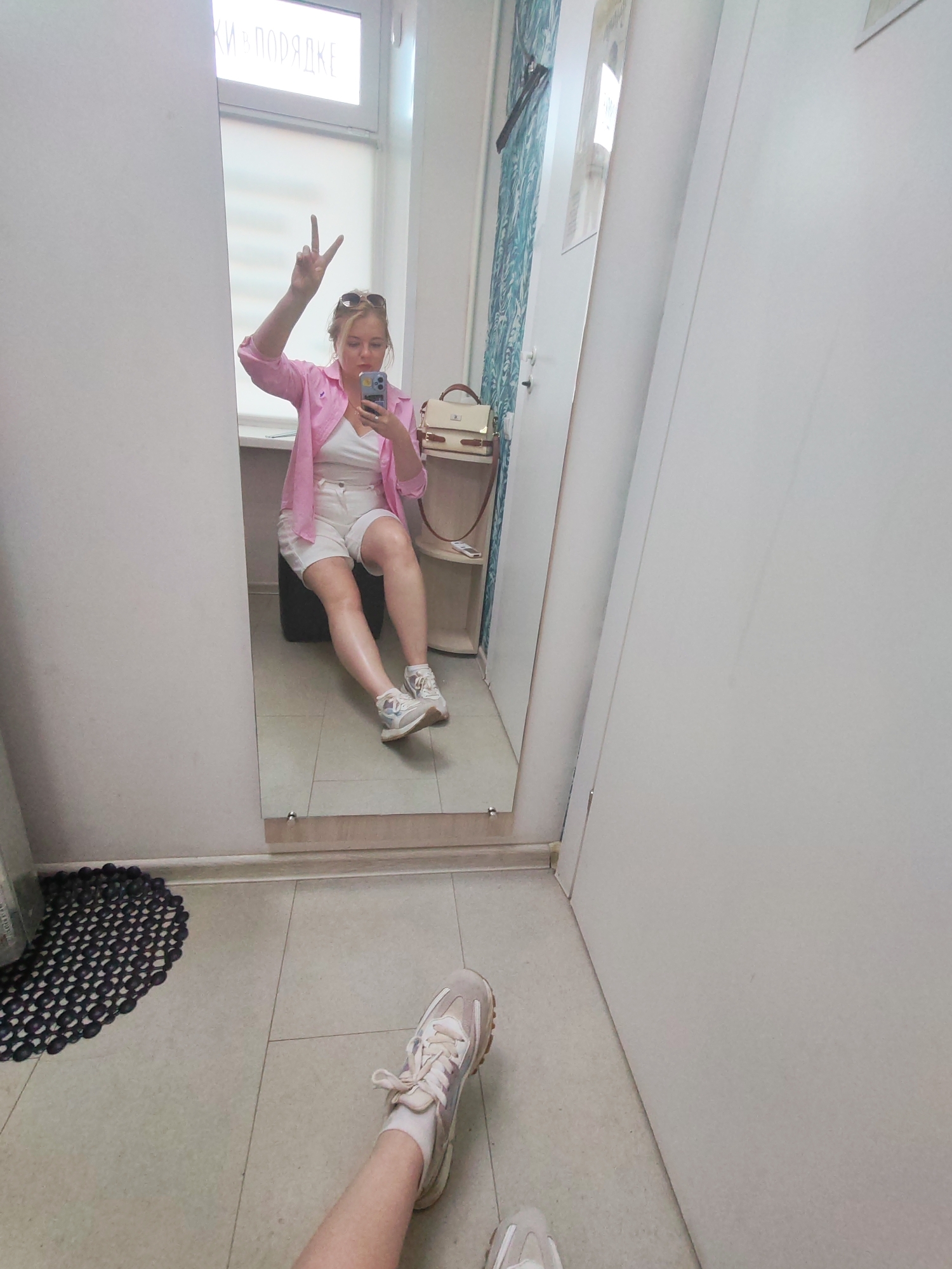 A full-length photograph shows a woman standing in front of a mirror, presumably in a dressing room. The location is Izhevsk, Russia, based on a subtle inscription visible in the reflection. The woman in the reflection is seated on a black object, wearing a white top, white shorts, a pink shirt, sunglasses on her head, and sneakers. She is holding a smartphone, appearing to take a selfie, and holds up a peace sign with her hand. Adjacent to her is a small table or shelf with a purse on it. In the foreground is a leg with a white sock and sneaker, and a second sneaker is visible. A black rubber mat with a circular pattern is positioned to the left of the leg and foot. The environment appears to be a room with a light-colored floor and white walls. A door is slightly ajar on the right side of the image.