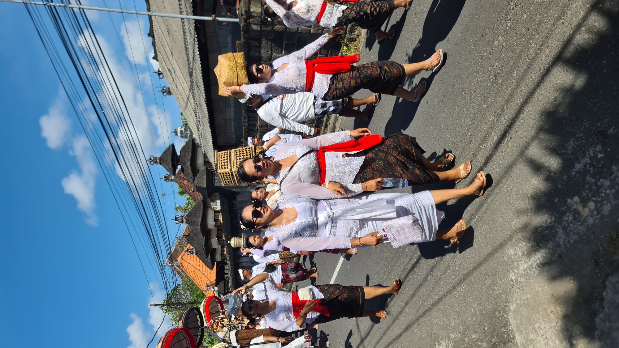 In Denpasar, Indonesia, under a clear blue sky with scattered clouds, a procession moves down a paved street. The participants, primarily women dressed in traditional white kebayas and sarongs, are adorned with red sashes. Some women carry woven baskets or offerings on their heads. The street is lined with stone walls and traditional Balinese architecture, characterized by multi-tiered roofs topped with decorative elements. Electrical wires are visible overhead. The scene captures a cultural event or religious procession taking place outdoors in a tropical urban environment.
