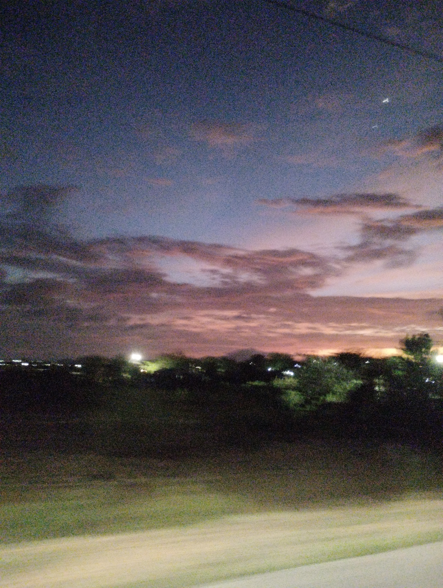 The image depicts a twilight scene, likely a rural or semi-rural area on the outskirts of La Guardia, Venezuela. The sky is a gradient of deep blue transitioning to hues of pink and orange, indicating sunset or sunrise. Wispy clouds are scattered across the sky, some illuminated by the fading light. In the distance, silhouetted trees and low-lying vegetation form a dark horizon. Scattered lights from what appear to be buildings or streetlights are visible through the trees, suggesting a human settlement. The foreground is a dark, blurred area, possibly a field or roadside, with a lighter, indistinct strip that could be a road or pathway. The image quality is grainy, characteristic of low-light photography. No people or specific landmarks identifiable as being in La Guardia, Venezuela, are visible.