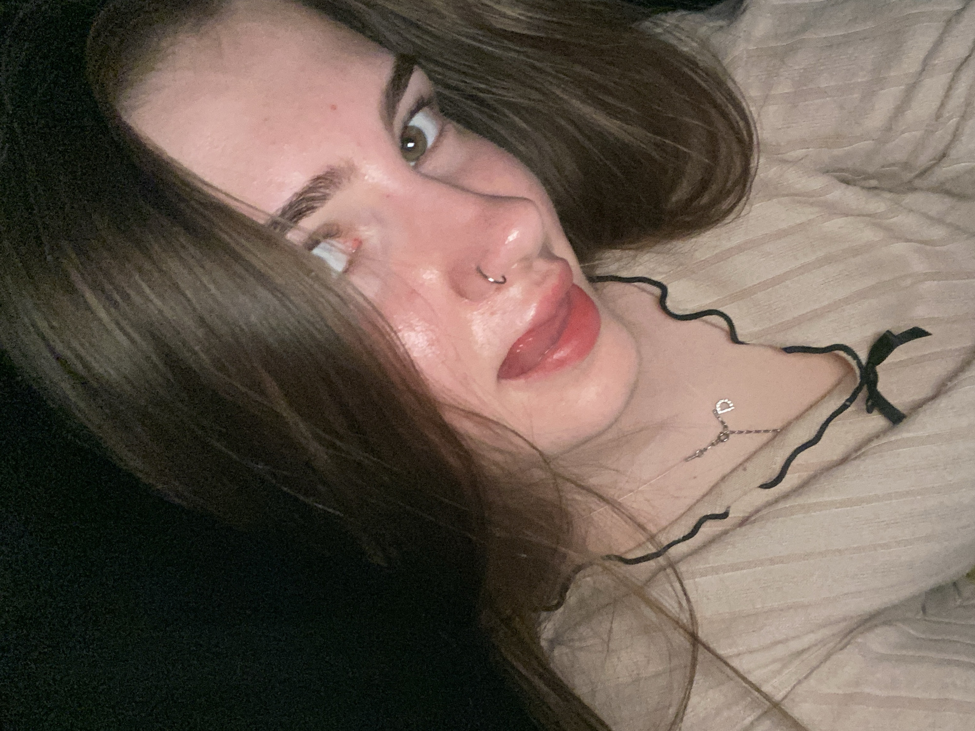 The image is a close-up, low-angle selfie of a young woman with long, brown hair. She is looking towards the camera with her head tilted. Her eyes are light brown, and she has a nose ring. Her lips are painted red and slightly parted. She is wearing a beige ribbed top with a black scalloped trim and a small black bow at the neckline. A silver necklace with a 'D' charm is visible around her neck. The background is dark and out of focus. There is no indication of the location being Дьяково, Russia.