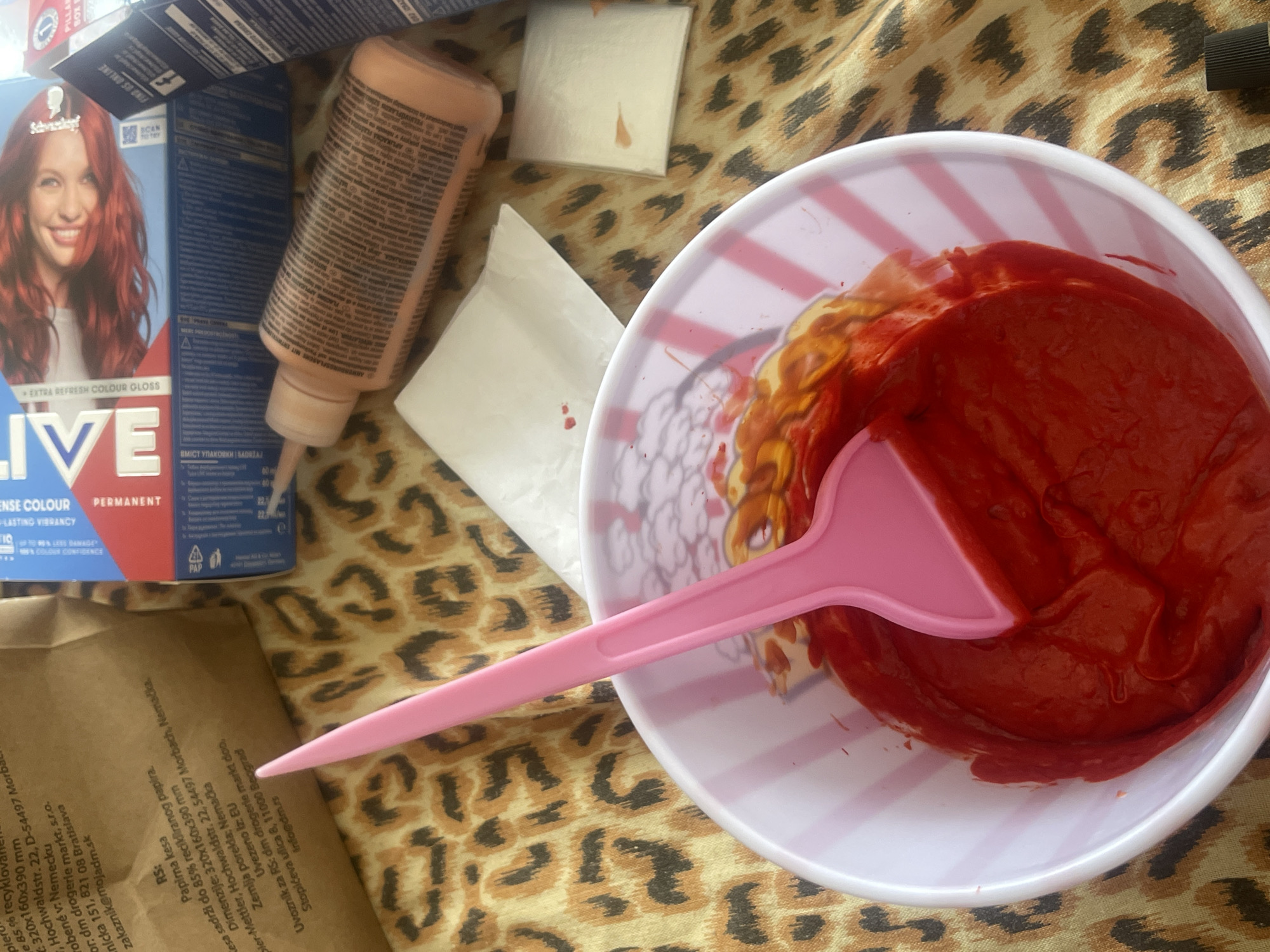 The image shows a close-up view of a plastic bowl filled with a thick, red substance, likely hair dye, being stirred with a pink plastic spatula. The bowl has pink and white stripes. Behind the bowl, a leopard-print fabric is visible, serving as a backdrop. Several product boxes and a small plastic bottle with an orange-colored liquid are scattered around the bowl. There are also small white squares of paper, one with a red stain. The overall scene suggests an at-home hair coloring process. The image does not provide any specific information about the city of Varna, Bulgaria.