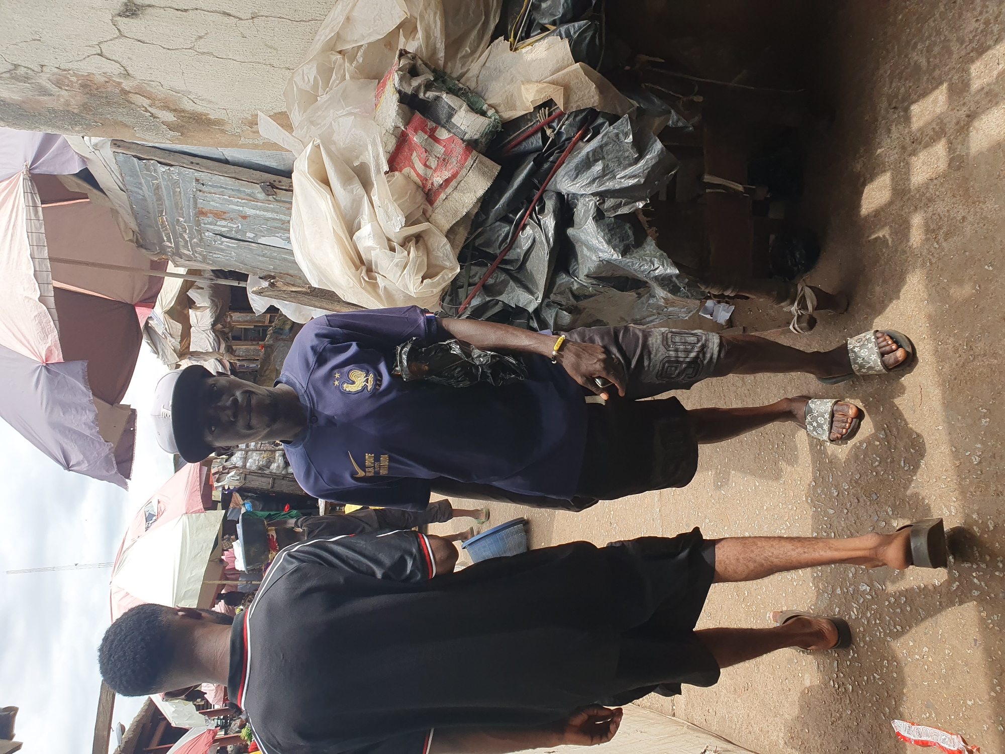 The image shows an outdoor scene in Lokoja, Nigeria. Two men stand on a concrete surface, likely a street or sidewalk. The man in the foreground wears a black shirt with white and red trim. Behind him, the other man wears a dark blue t-shirt with a white cap. They both wear shorts and sandals. In the background, there are several market umbrellas, and what appears to be a building or stall with materials piled on top, including white and black plastic sheeting. The overall impression is of a bustling outdoor market setting.