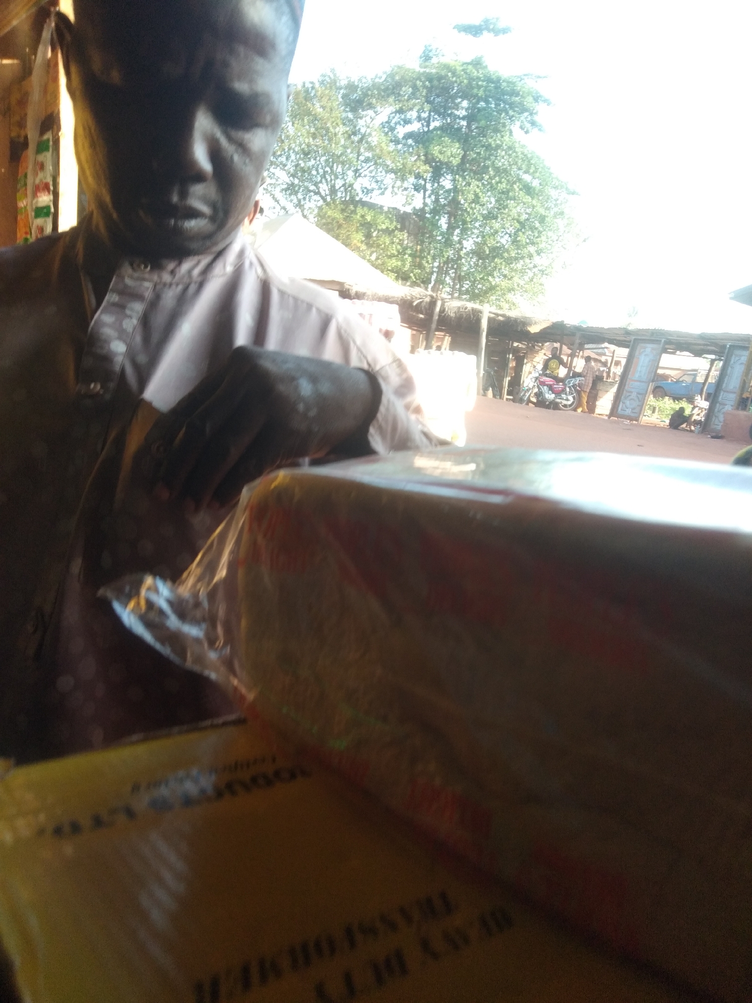 A man stands indoors near a window in Ejule, Nigeria, holding a packaged item. He wears a pink shirt with polka dots and looks down at the package. Through the window, an outdoor scene is visible: a street with people, motorcycles, and simple roadside structures under a partially obscured tree.