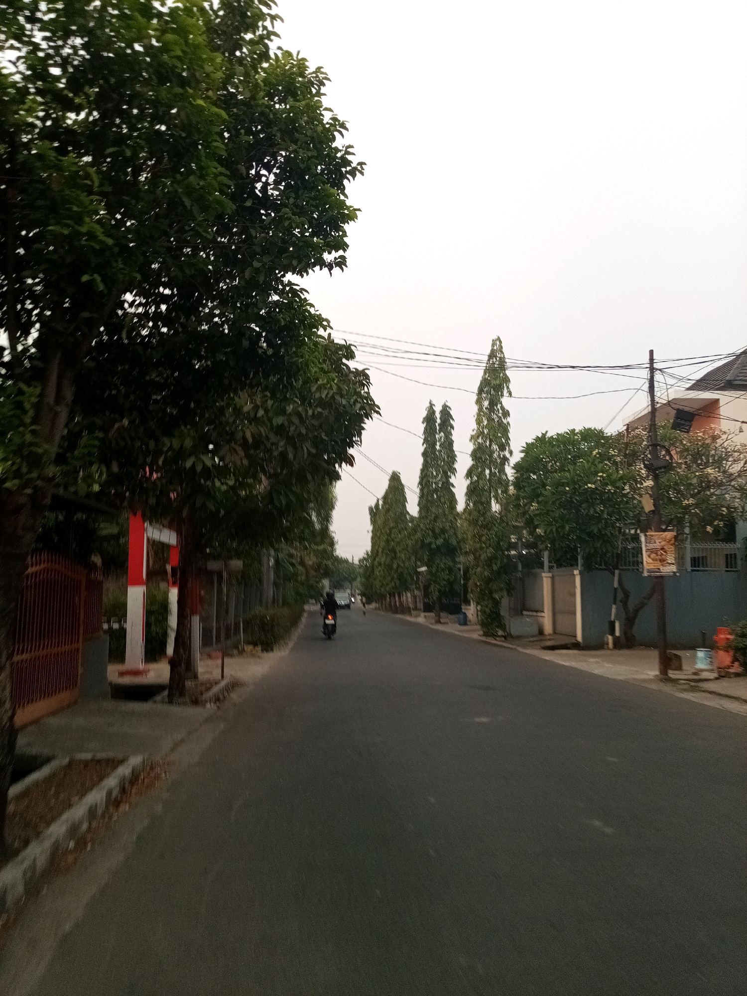 An eye-level shot shows a residential street in Bekasi, Indonesia. The street is asphalt and curves to the right. On the left side of the road, a tall, leafy tree partially obscures a red and white striped pole and a brown metal fence. Further down the street, a person on a motorcycle is riding away from the camera. On the right side of the street, there are several tall, slender trees lining a wall. A building with a dark roof and cream-colored walls is visible behind the trees. The sky is overcast and pale gray.