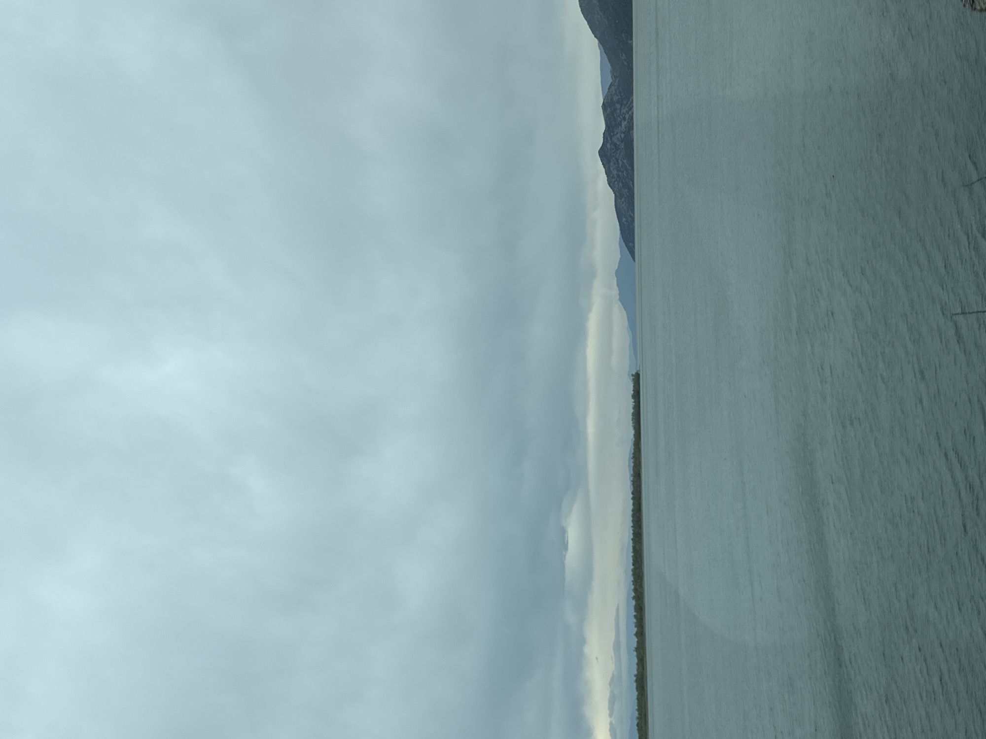 The image shows a body of water, likely a lake or fjord, under a cloudy sky. A tree-lined shore separates the water from the distant mountains. The water is calm and reflects the sky. Kruševica, Montenegro, is not visibly identifiable in this image.