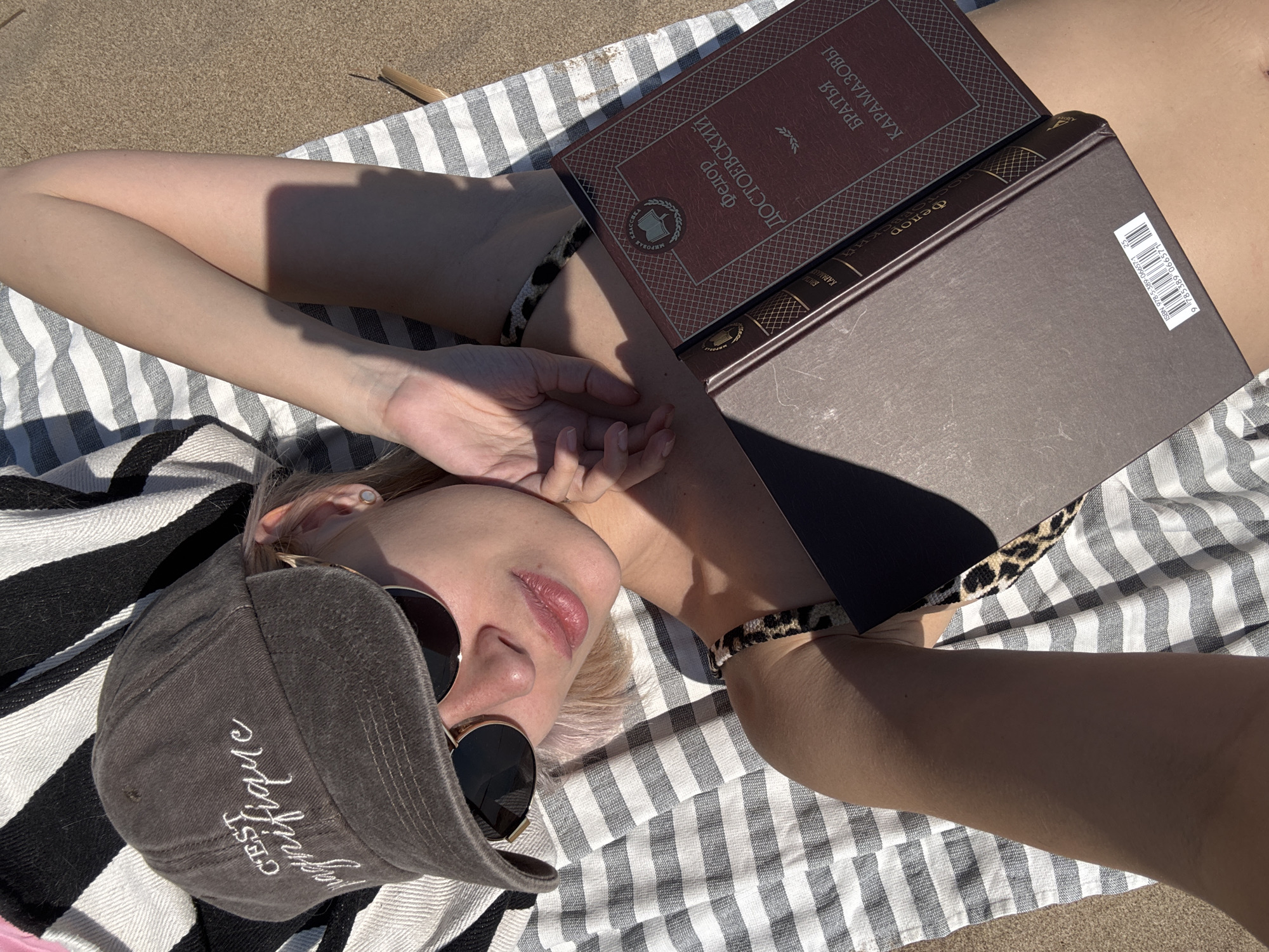 A woman lies on a striped towel on the beach. Her face is partially covered by a gray baseball cap and sunglasses. She is holding a copy of "The Brothers Karamazov" by Fyodor Dostoevsky in Russian on her chest. The ISBN number on the book is 978-5-389-06657-1. The location is presumably a beach, but the exact location in the city of Saratov, Russia, cannot be determined from the image.