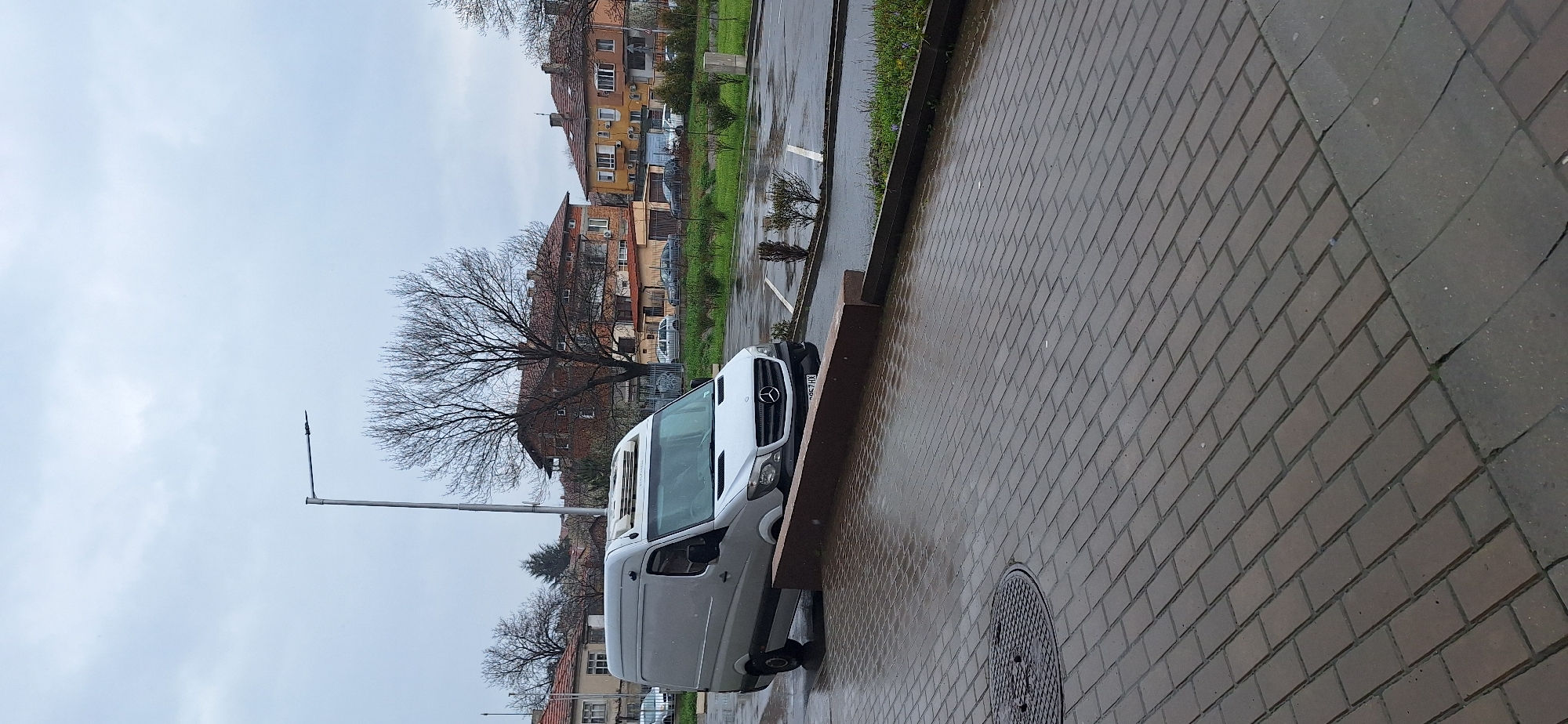 The image depicts an urban scene in Burgas, Bulgaria. The ground is covered with wet, reddish-brown paving stones. A light-colored Mercedes-Benz van is parked alongside the curb, partially obscuring the curb line. A street lamp stands nearby. Beyond the van, there are several multi-story buildings with pitched roofs, interspersed with bare trees and some greenery. The sky is overcast and gray. A manhole cover is visible in the foreground.