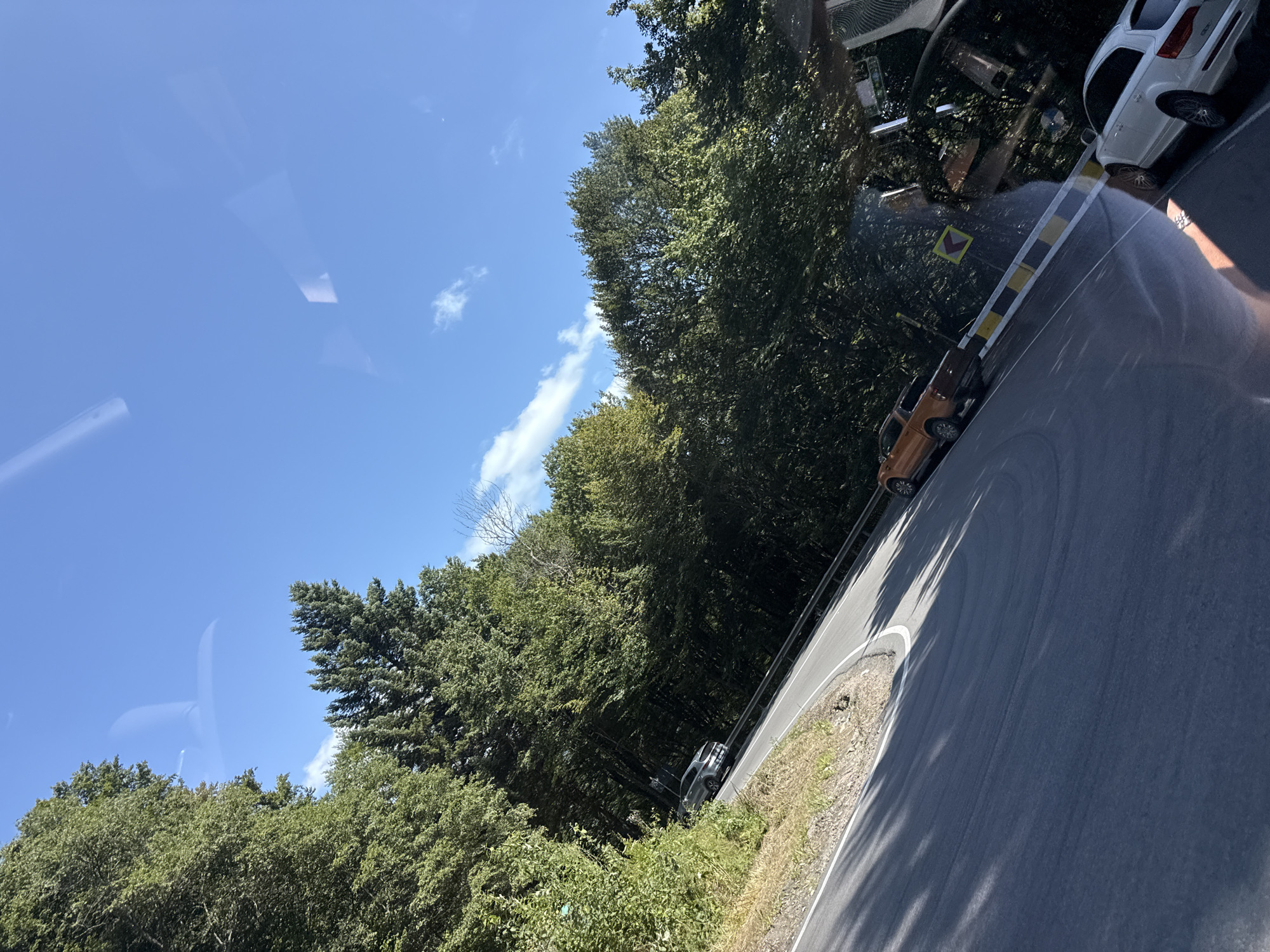 The image depicts a winding road through a forest, likely in the Oituz, Romania area. Several vehicles are visible on the road. In the foreground, a white SUV is parked on the right side of the road. Further up the road, an orange pickup truck is driving. To the left, a silver SUV is also visible on the road. The road is flanked by dense green trees. The sky is blue with a few scattered clouds. Reflections are present on the surface from which the photo was taken, suggesting it was captured from inside a vehicle.
