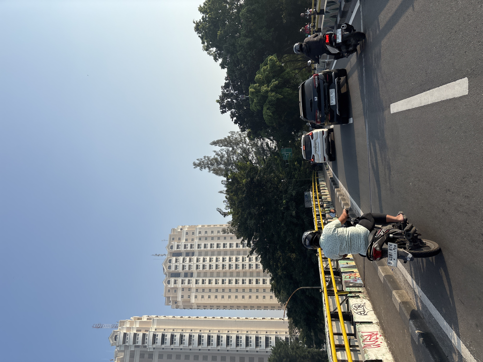 This is a photograph taken on a sunny day in West Jakarta, Indonesia, likely in the late morning or early afternoon, given the bright sunlight casting distinct shadows. The image captures a scene of urban traffic on an elevated road or bridge.
The primary subjects are vehicles and people on motorcycles, typical of Southeast Asian cityscapes. Several cars are visible in traffic, including a black SUV and a white hatchback. The foreground is dominated by a black motorcycle with a rider wearing a light-colored patterned shirt and dark pants, along with a helmet. The motorcycle's license plate reads "B 4981 BTK". Another motorcycle is further up the road, also with a rider wearing a helmet. In the background, more vehicles and trees can be seen.
The setting is a multi-lane road with white lane markings. A yellow railing and concrete barriers line the side of the road, with some graffiti visible on the barriers. Beyond the road, tall, modern residential buildings with many windows are prominent in the cityscape, indicating an urban environment. Lush green trees are also abundant, providing a contrast to the concrete structures. The sky is clear and a light blue, suggesting good weather conditions. The overall impression is of a busy urban commute on a bright day.