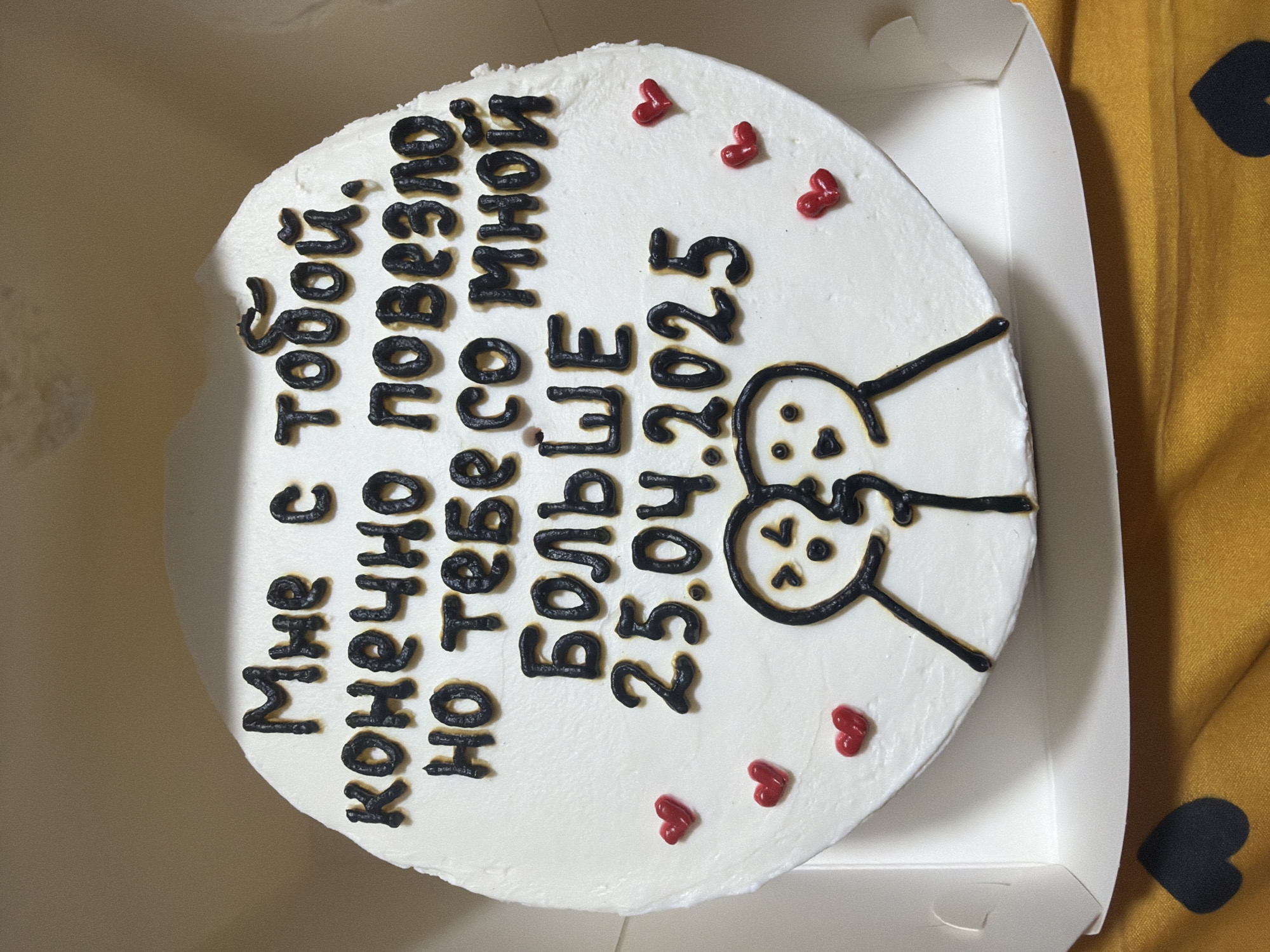A close-up, overhead shot shows a round cake inside a white cardboard box, likely taken indoors. The cake is decorated with white frosting and black icing. Black icing is used to write a message, which is presumably in Russian (Мне с тобою, конечно, повезло, но тебе еще больше со мной. 25.04.2025). The icing is also used to draw a simple image of two cartoon figures touching heads. Several small, red, heart-shaped decorations are scattered around the cake. The box is sitting on a mustard yellow fabric with black heart patterns. No people are visible. Location is undetermined.