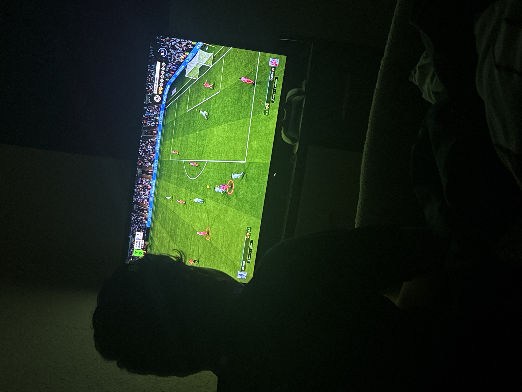 A person is lying down in a darkened room, facing a television. The television displays a soccer video game, with a view of the green field, players, and goal. The scoreboard indicates the game is between teams "CAM" and "BMA," with the score tied at 1-1. Text on the screen also includes "EA SPORTS" and "999.XING." Due to the darkness and angle, specific facial features of the person are indistinguishable. The environment appears to be a residential setting. The location, Welshpool, United Kingdom, cannot be ascertained from the image alone.