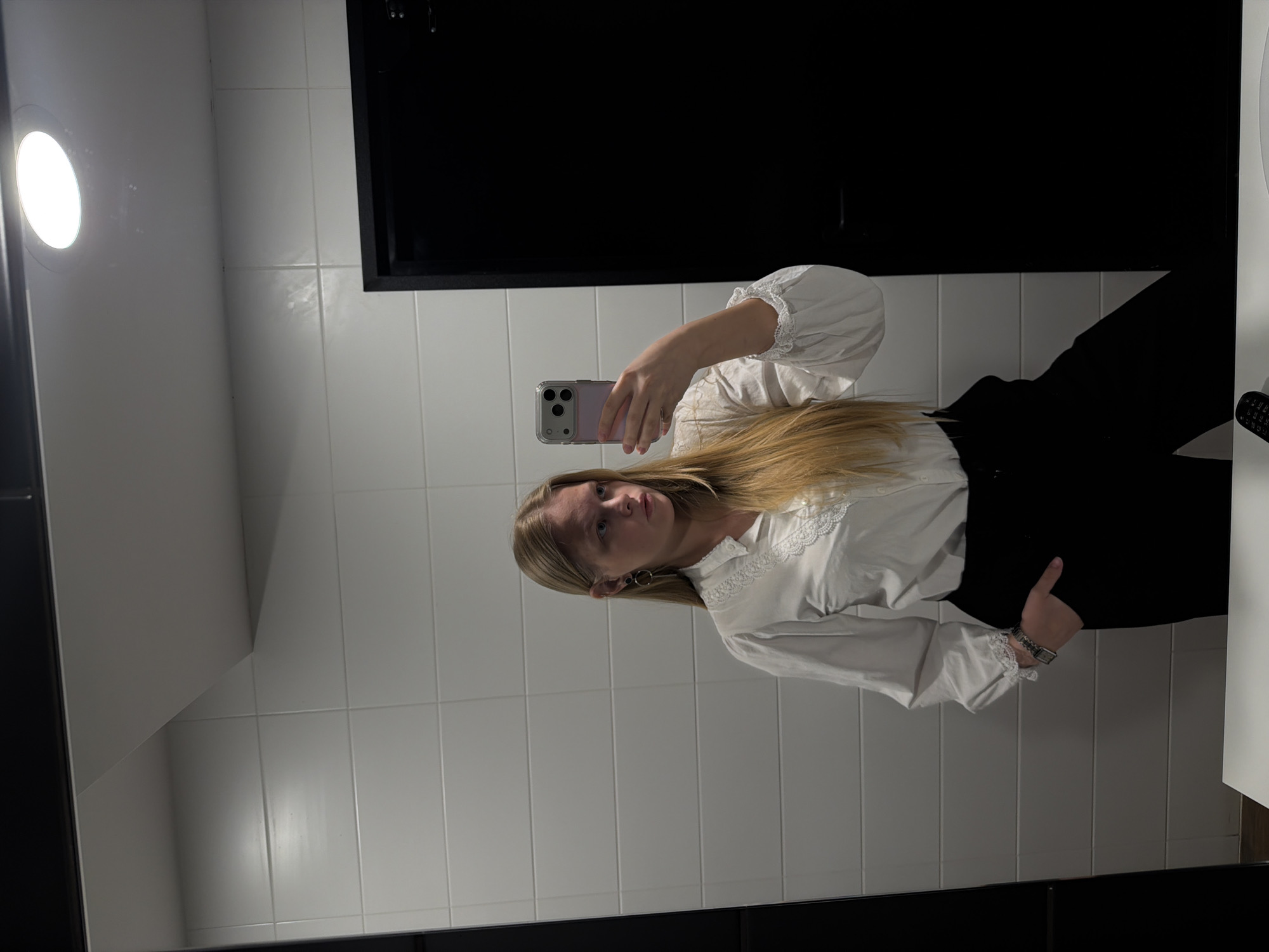 A young woman with long, straight blonde hair and a neutral expression is captured in a mirror selfie, looking directly at the camera. She wears a white long-sleeved blouse featuring a ruffled or lace-trimmed collar and cuffs, paired with dark trousers or a skirt. A silver-colored watch is visible on her left wrist, and she has circular silver earrings. She holds a light purple/pink smartphone with a clear case in her right hand, using it to take the photograph.
The setting appears to be a modern indoor space, likely a bathroom or restroom, characterized by white rectangular tiled walls. A prominent, bright circular light fixture is visible in the reflection on the upper left. A large, dark rectangular area dominates the background behind the woman, possibly a dark wall, a dark mirror frame, or a window. There are no visible texts in the image. The photo provides no specific time of day or weather cues, as it is taken indoors under artificial lighting. The overall impression is a posed, casual mirror selfie.