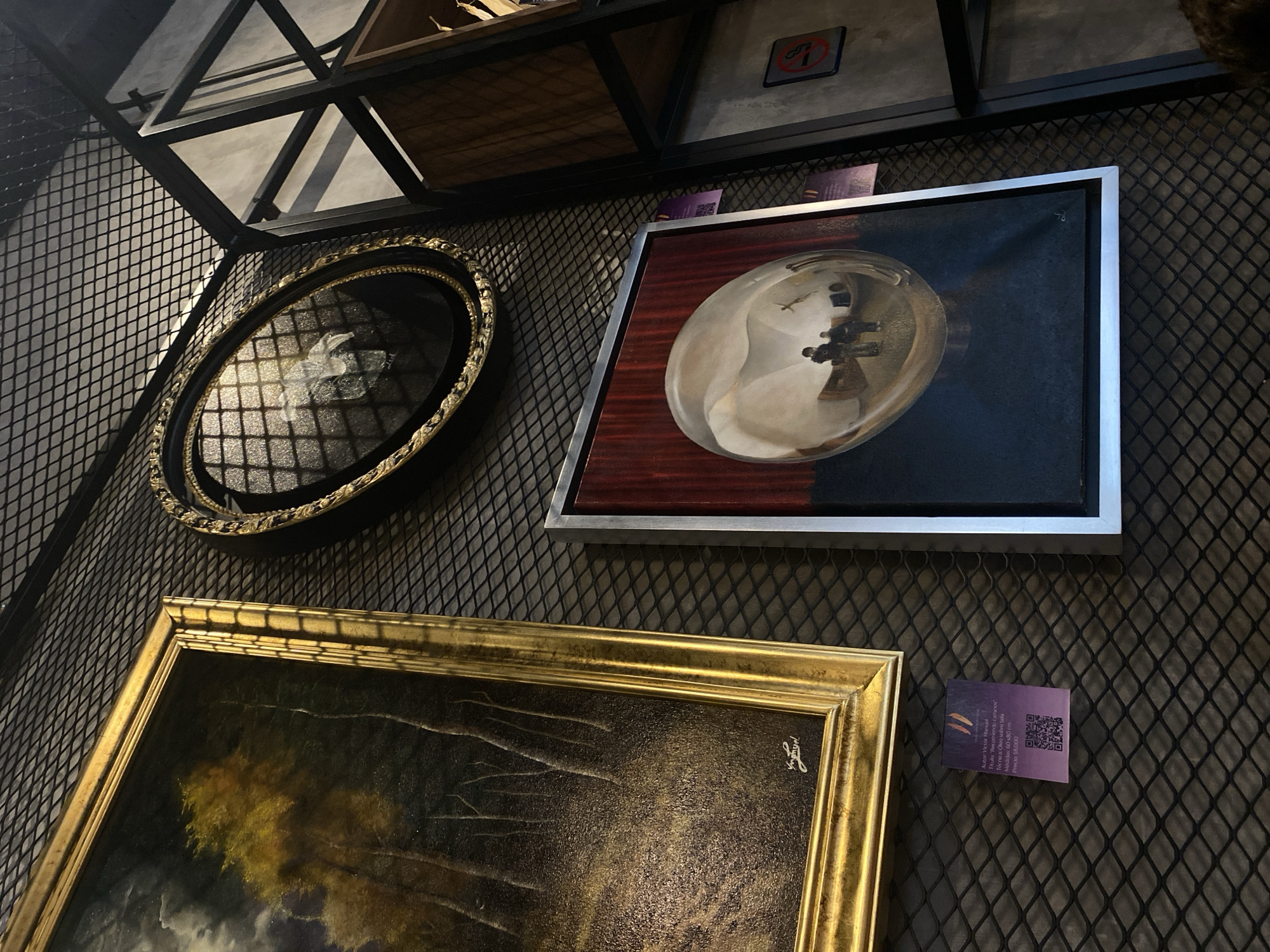 The image shows a display of three framed artworks on a dark gray mesh wall, possibly in an art gallery or exhibition space. The top left artwork is an oval-shaped frame with an indiscernible image within. To its right is a rectangular framed artwork with a silvery, reflective central image against a dark background, bordered by a reddish-brown inner frame and a silver outer frame. Below these two, a larger rectangular framed artwork with a gold frame is visible, depicting what appears to be a landscape painting. A small purple sign with text and a QR code is affixed to the wall near the gold-framed painting. Above the artworks, a black metal structure forms a shelf or display rack, partially obscuring the background. A second similar sign is visible between the oval frame and the rectangular artwork with the reflective image.