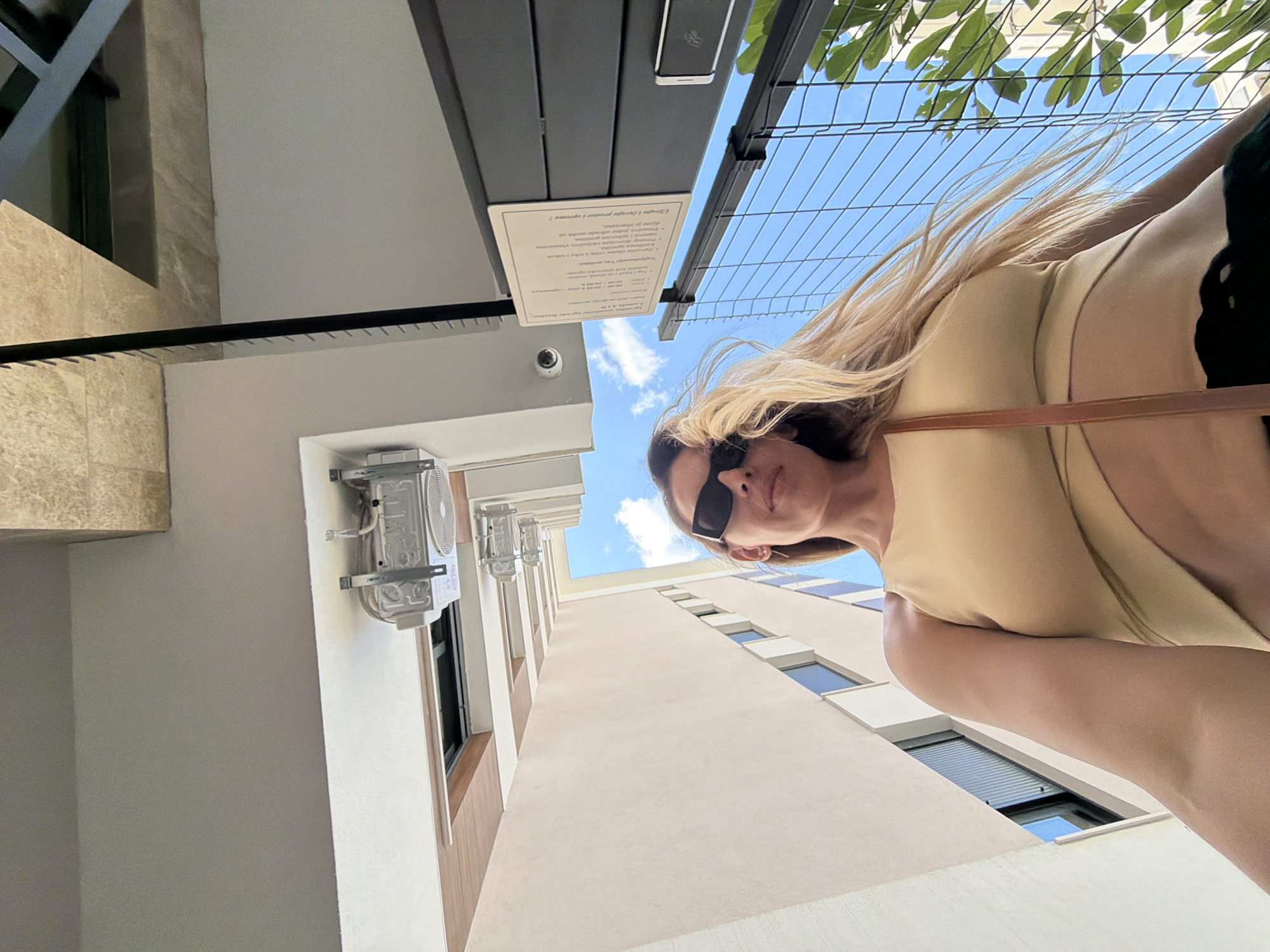 The image, taken in Budva, Montenegro, shows a woman from a low-angle perspective. She is wearing sunglasses and a light-colored, possibly beige, crop top that wraps around her torso. Her blonde hair is blowing in the wind. Behind her, a tall, light-colored building with dark-framed windows stretches upwards. Above, a light grey ceiling meets a beige textured surface, with what appears to be an outdoor fan mounted on the ceiling. The background also includes a clear blue sky with some clouds, and a dark metal fence with greenery visible through it. A sign with text is attached to a dark metal structure on the right side of the image.