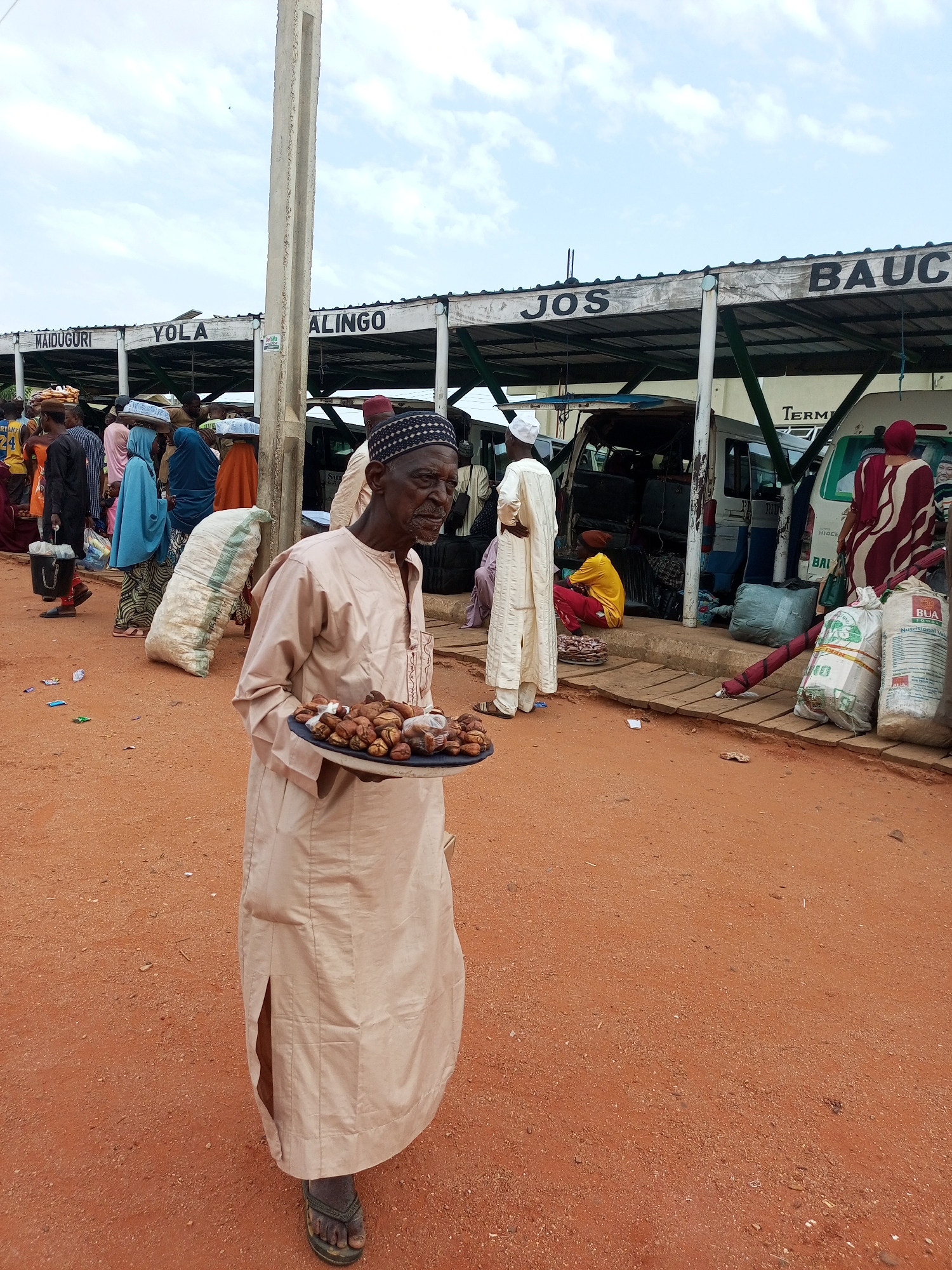 An elderly Black man in a tan robe and a black and white cap stands in the foreground of an outdoor market, holding a circular black tray filled with dried fruits or nuts. He is looking to his right. Behind him, a covered market stall is visible with signs reading "MAIDUGURI," "YOLA," "ALING," and "JOS," "BAUCH." Several people, some carrying items on their heads or in bags, are gathered under the stall. The ground is reddish-brown dirt, and there are several large bags and some vans parked in the background. The sky is partly cloudy.