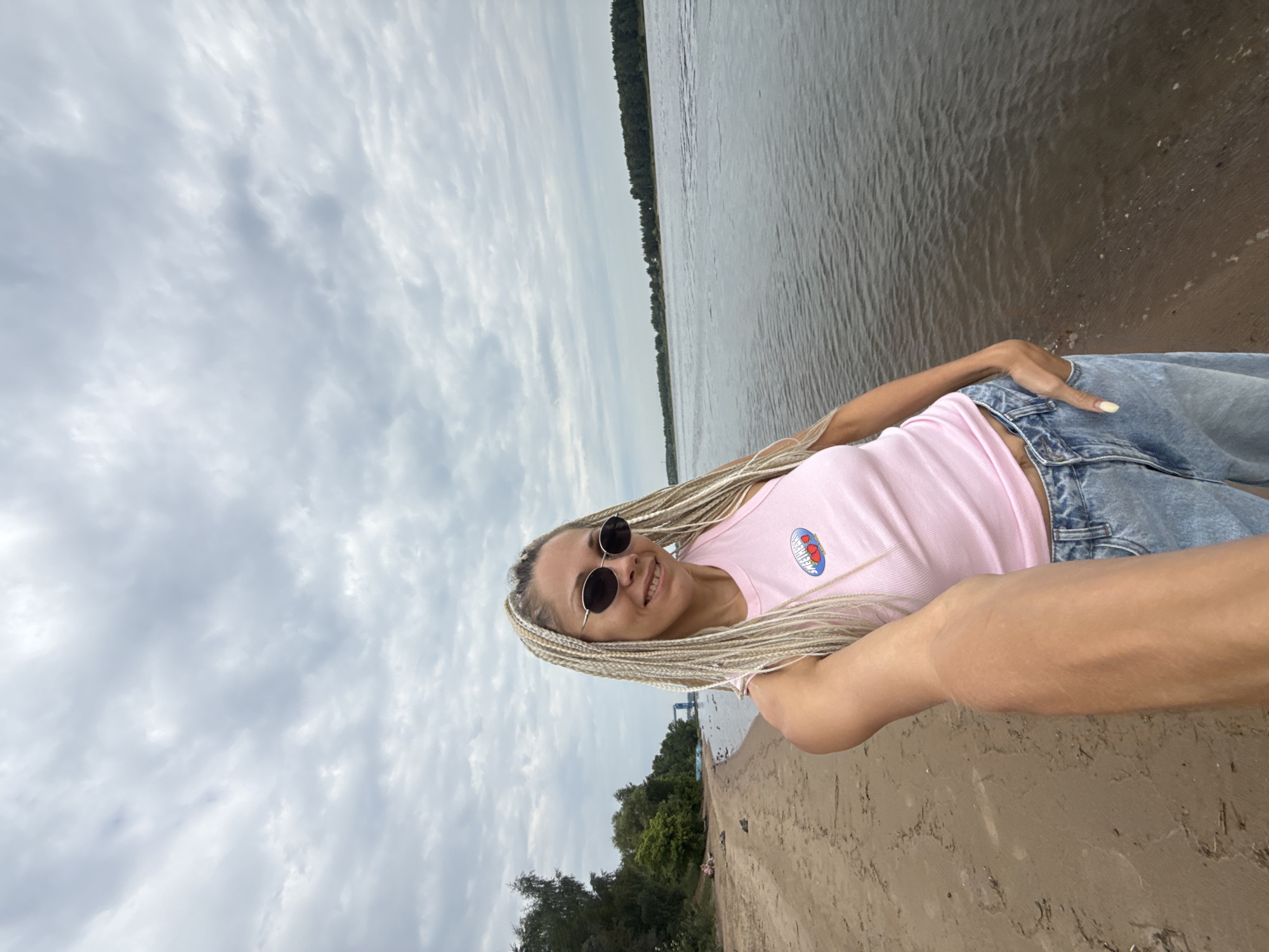A woman with blonde braided hair and sunglasses is taking a selfie on a sandy beach with a river in the background. She is wearing a pink tank top and blue jeans. The sky is overcast. The location is the city of Dumino, Russia.
