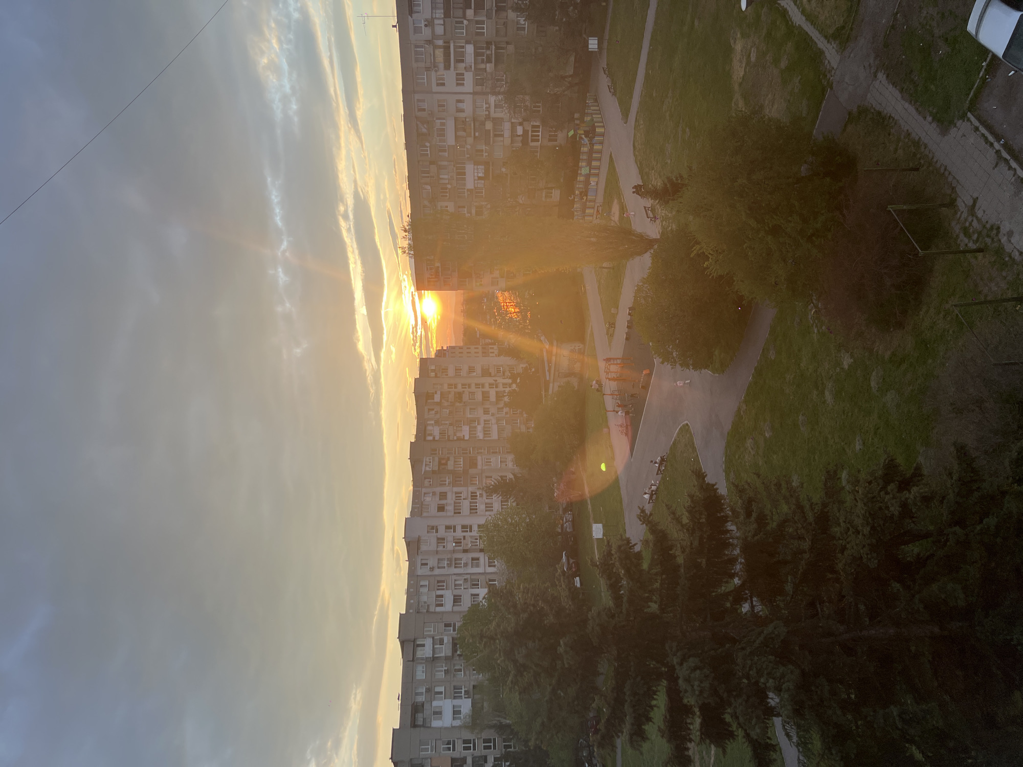The image captures a residential area in Sofia, Bulgaria, at sunset, viewed from an elevated perspective. The warm, golden light of the setting sun dominates the left side of the frame, casting a strong glow and sunbeams through a partly cloudy sky. Several multi-story apartment blocks, typical of Eastern European architecture, form the backdrop and mid-ground. These buildings feature numerous windows and balconies, with some exhibiting air conditioning units.
Below the buildings, expansive green spaces are visible, interspersed with trees, both deciduous and coniferous, and winding paved pathways. A colorful outdoor play or seating area can be discerned near one of the apartment blocks. A few indistinct figures, likely people, are scattered on benches or walking on the paths, enjoying the evening. A white car is partially visible on a road or parking area on the right edge. A thin power line stretches across the upper left sky, and an antenna is visible on a rooftop. The scene conveys a tranquil end to the day in an urban environment. No discernible text is visible.