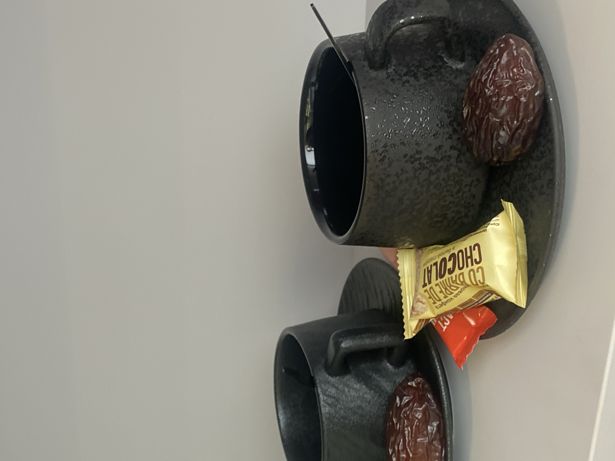 Two black cups and saucers are stacked on top of each other, angled to the right, with a date and a wrapped chocolate candy bar on each saucer. The scene appears to be indoors, against a light-colored wall. Votkinsk, Russia is not depicted in the image.