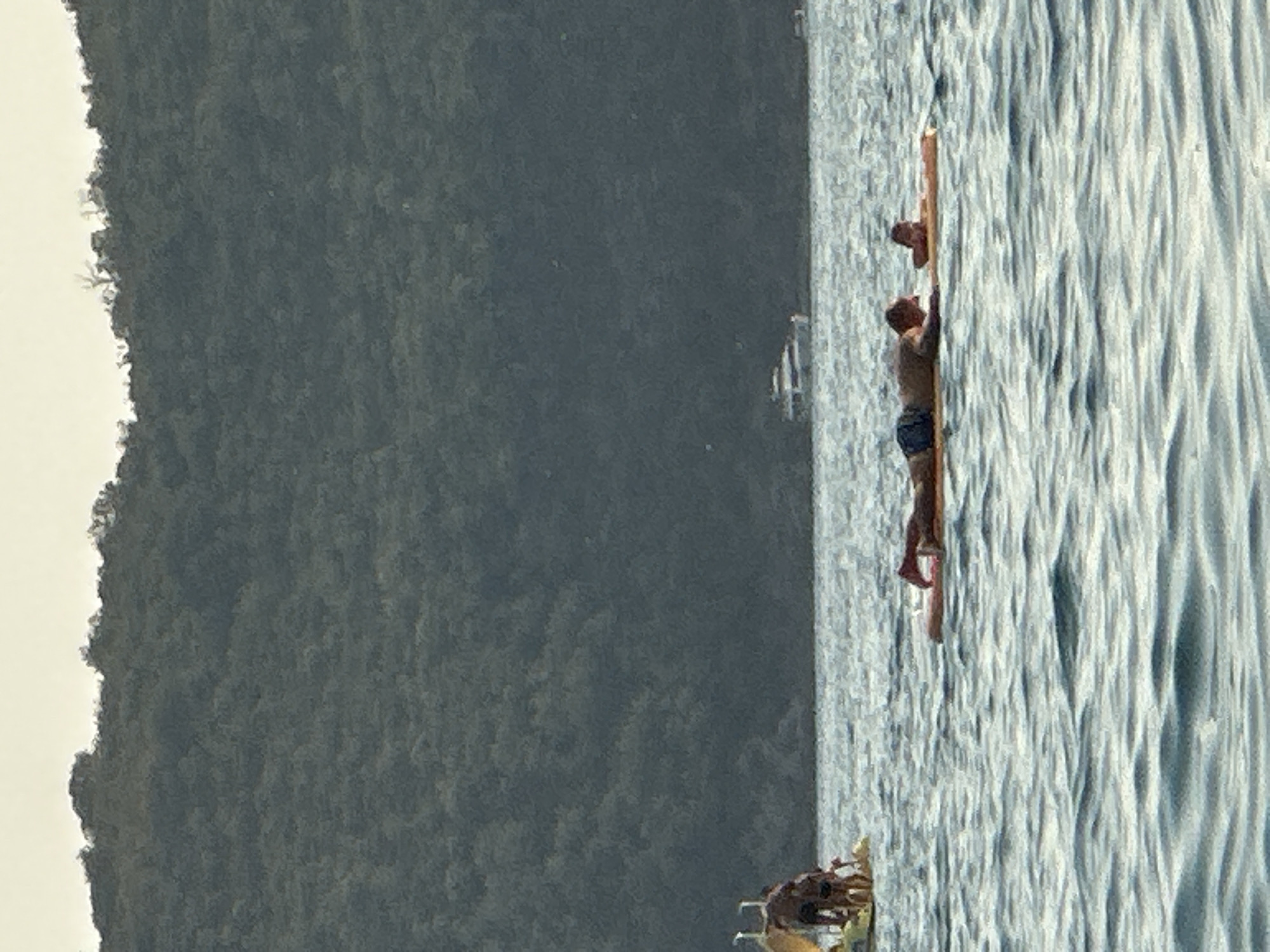 Two people are on a raft in the water, with a man in front and a child behind him. The man is lying on his stomach, facing left, with his arms outstretched. The child is also lying on their stomach, facing right. The raft is orange and appears to be made of foam. In the background, there is a large hill covered in trees. A white boat is visible in the distance, and another smaller boat is further to the right. The water is calm with small ripples. In the far left of the image, there is a yellow structure with people standing on it, seemingly in shallow water. This scene is likely located in the waters near Boreti, Montenegro, given the presence of the distinctive hills and the type of coastline visible.