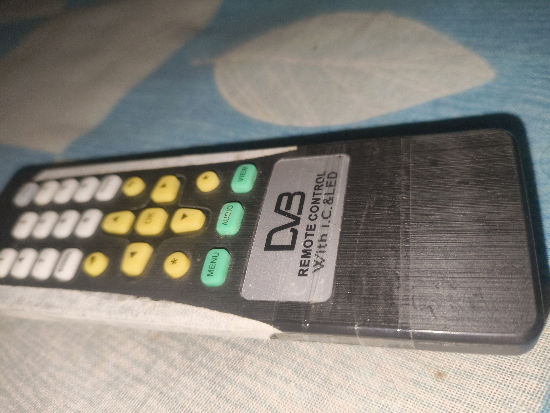The image shows a close-up, high-angle shot of a black DVB remote control lying on a textured blue and white patterned surface, possibly a bedsheet or tablecloth. The remote is positioned diagonally, with the top left corner closer to the viewer. The remote has a variety of buttons, including white numbered buttons, yellow directional and function buttons (OK, Play/Pause, Skip Forward, Skip Backward), and green function buttons (AUDIO, VIEW, MENU). A silver sticker on the right side of the remote has "DVB REMOTE CONTROL With I.C. & LED" printed on it. The surface the remote is on has a slightly worn and dusty appearance, with some faint stains visible. The lighting appears to be artificial and casts shadows, indicating an indoor setting. There are no people or any other discernible objects in the frame. The image does not contain any information that would allow for identification of its location in Khillupur, India.
