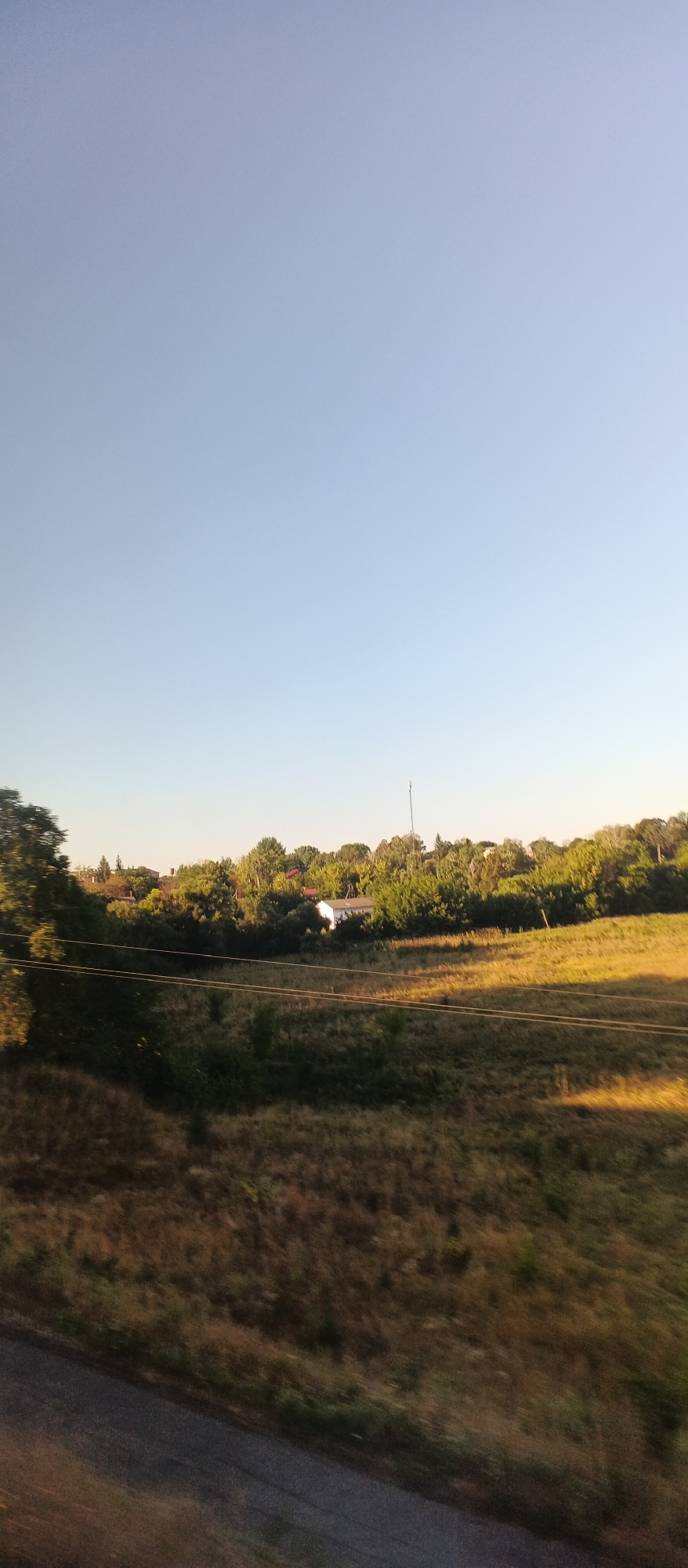 The image depicts a rural landscape under a clear, pale blue sky. In the foreground, a dark, asphalt road runs diagonally from the bottom left corner towards the middle right. Adjacent to the road, a field of dry, golden-brown grass stretches across the lower half of the image, with hints of green vegetation interspersed. A slight blur suggests motion, possibly from a vehicle.

Further into the midground, the terrain slopes upwards, covered in a mix of dry grass and darker green shrubbery. Several thin, horizontal wires, likely power lines, are visible stretched across this area. Behind the wires, a line of dense trees forms a verdant backdrop, with varying shades of green indicating different species and growth.

Within the treeline, nestled amongst the foliage, are several buildings with red roofs, and a prominent white rectangular structure. A tall, slender tower, possibly a communication mast, rises above the trees on the right side of the frame. The scene suggests a warm, possibly late afternoon or early morning light, casting long shadows across the field. The overall impression is one of a quiet, undeveloped area, likely on the outskirts of a settlement. The location is identified as Khlystunivka, Ukraine.