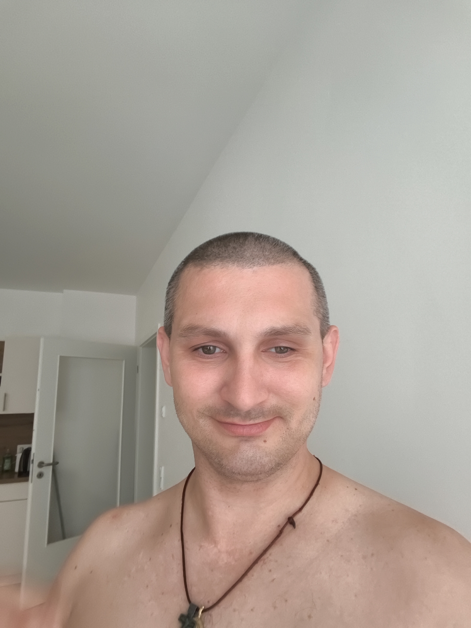 A man with a buzz cut is looking at the camera and smiling. He is wearing a necklace. The background is a plain white wall. The man's shoulders and upper chest are visible. A door and a cabinet are visible in the background to the left of the man. The image was taken indoors in Wolgast, Germany.