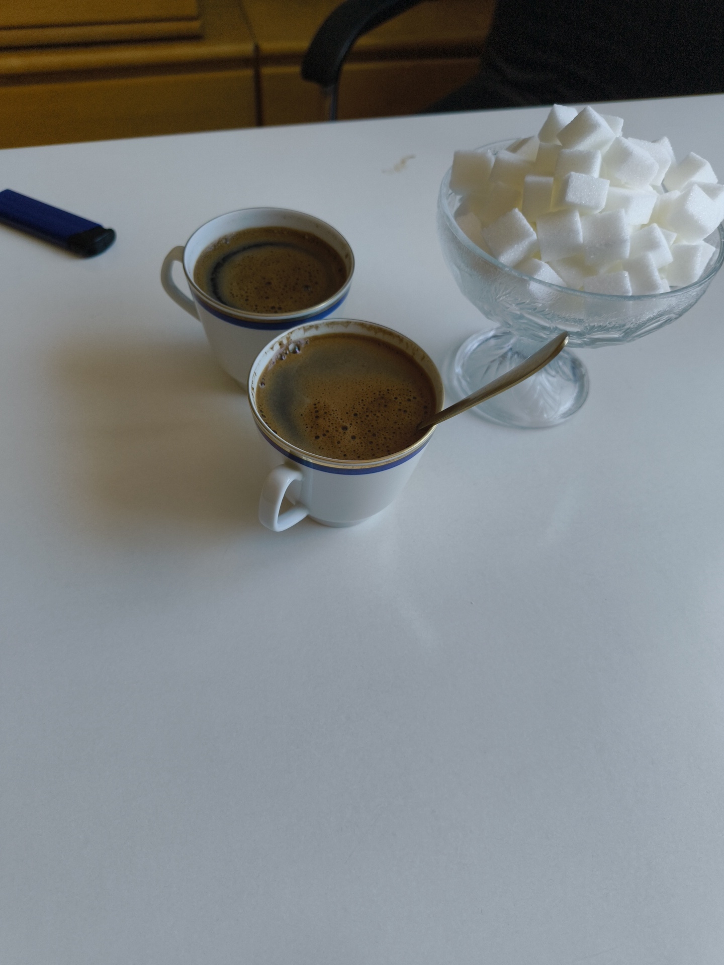 A white table in Hardthausen am Kocher, Germany, holds two white coffee cups filled with dark coffee, a glass dish overflowing with sugar cubes, and a spoon resting in one of the cups. To the left of the cups is a blue object, possibly a pen or lighter. The background shows a wooden surface and a glimpse of a dark chair. The scene is illuminated by artificial light, casting soft shadows on the table.