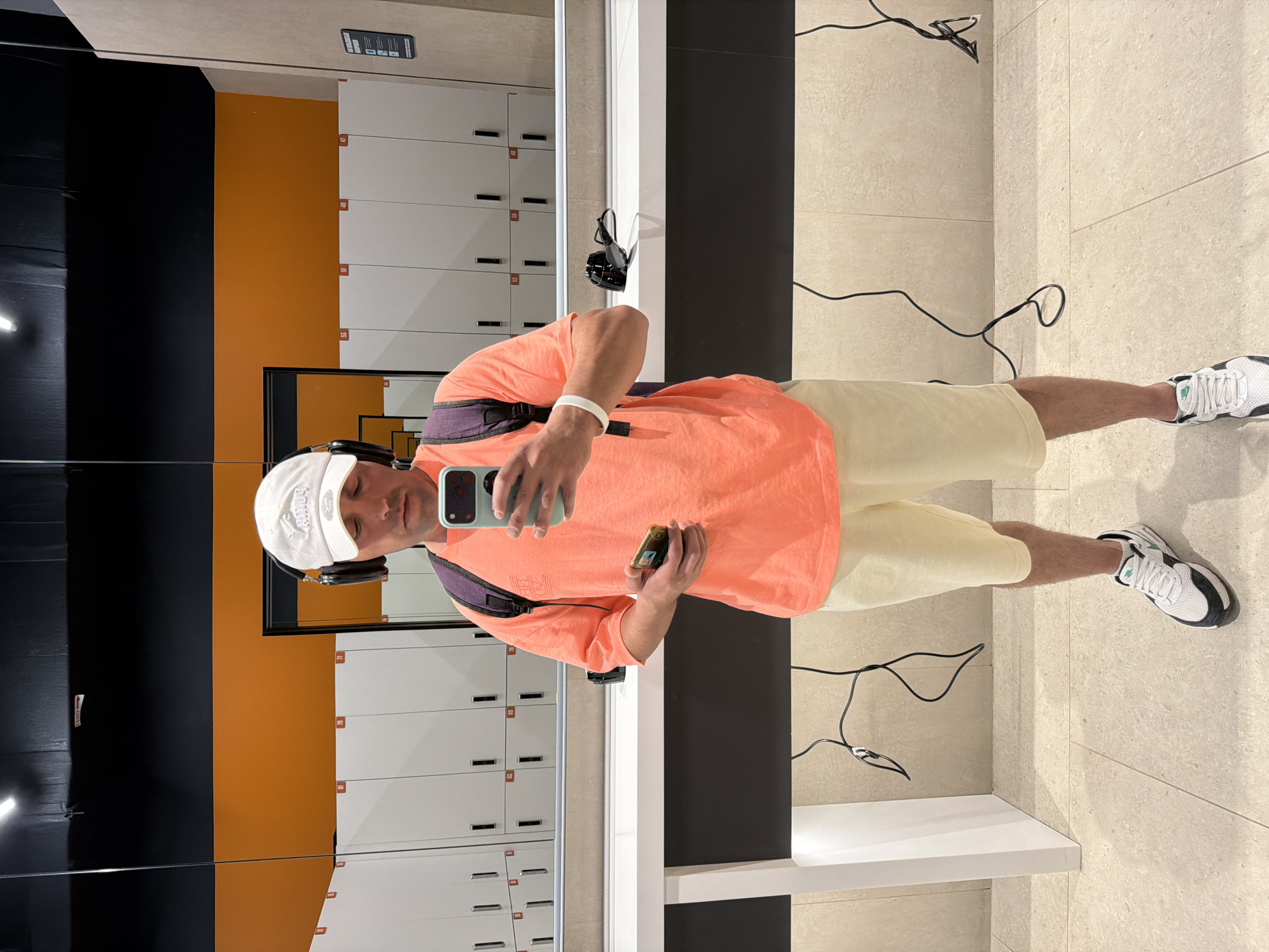 An indoor mirror selfie captures a man standing in what appears to be a modern locker room or changing facility in Krasnoyarsk, Russia. The image is rotated 90 degrees clockwise. The man, seen in his reflection, is wearing a white baseball cap, black over-ear headphones, an orange short-sleeved t-shirt, light yellow shorts, and white and black sneakers with green accents. He has a white wristband on his left arm and a small black shoulder bag worn across his chest. He holds a mint green-cased smartphone in his left hand, pointing it towards the mirror to take the picture, and a small, dark electronic device (possibly a vape) in his right hand.
The background features white lockers with red numbered plates, visible numbers include 47, 48, 49, 50, 51, and 52. The walls behind the lockers are sectioned into black and vibrant orange panels. The floor is light-colored tiled. Black cables are visible on the floor, suggesting charging stations or equipment connections. Notable visible text includes an "EXIT" sign with an arrow on a black wall and a "No Smoking" icon with other symbols on a black sign above the lockers. The text on his cap is mirrored and stylized. The setting is brightly lit, indicative of an indoor public facility.