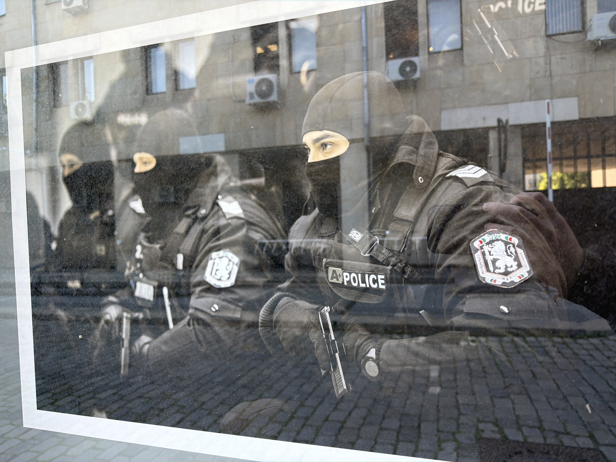 The image is a photograph, taken through a window, of a black and white photograph. The black and white photograph depicts three armed individuals wearing black balaclavas and dark uniforms with "Police" insignias. The individual closest to the viewer is holding a handgun and has a patch on their sleeve featuring a lion emblem surrounded by the colors of the Bulgarian flag. The backdrop visible through the window reveals an exterior building with air conditioning units and the reflection of the word "Police" in reverse, suggesting the photograph is displayed at a police station. The cobblestone street visible as a reflection in the glass suggests the location is Sofia, Bulgaria.