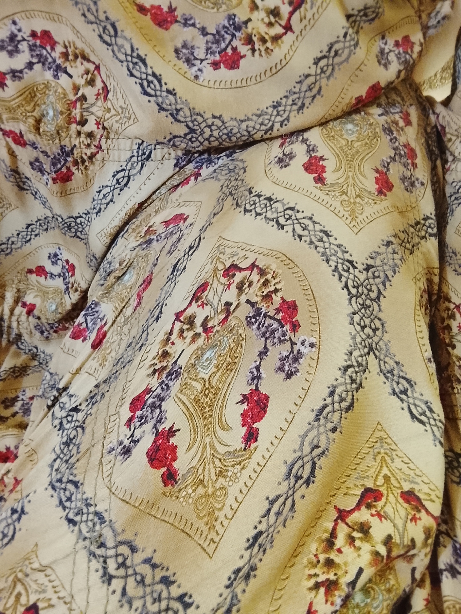 The image presents a close-up view of a patterned fabric. The fabric has a light beige or tan background with a repeating design featuring stylized floral arrangements in red, purple, and brown hues, set within decorative cartouches outlined with a darker ornamental pattern. The fabric is draped and creased, obscuring the repetition of the design and creating a textured effect. The scene does not provide information about the location, city, people, subjects, objects, actions, interactions, and environment.