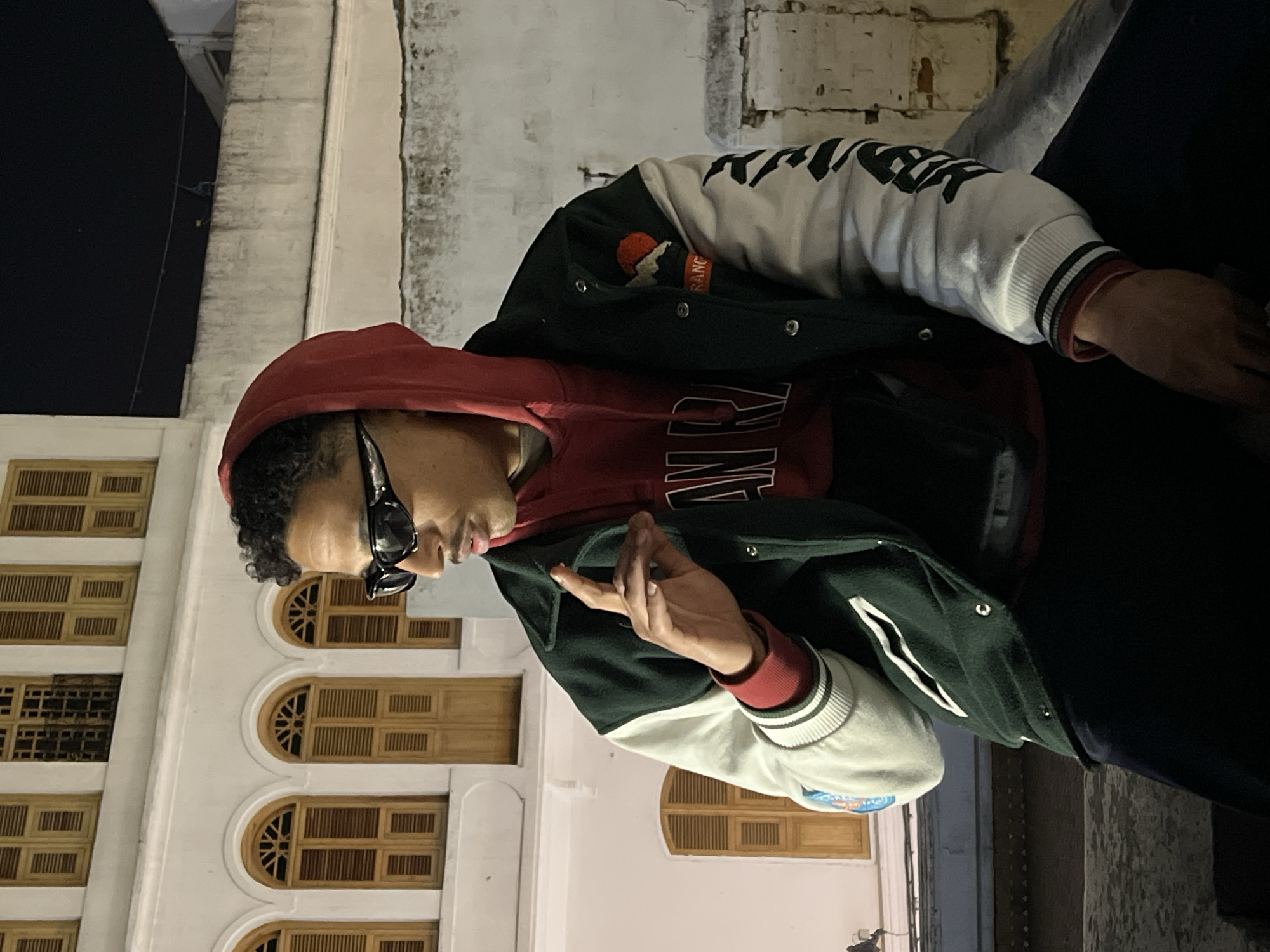 A young adult male is depicted in a profile shot against a nighttime urban background. He has dark, curly hair and a mustache, and is wearing black, narrow-frame sunglasses. His attire consists of a dark red hooded sweatshirt, with the hood pulled up, beneath a dark green varsity jacket featuring white sleeves. The jacket's right chest area displays a patch with a red sun or mountain motif, and its left sleeve has black script lettering. His right hand is raised to chest level, with fingers slightly curled as if gesturing. He appears to be seated or leaning on a low, light-colored concrete or stone barrier.
The background reveals a multi-story white building with distinct arched windows fitted with light wooden shutters, indicative of traditional Moroccan or Andalusian architecture. A less defined, weathered wall is visible further back to the right. The scene is illuminated by artificial light, casting some shadows. The architectural style and urban setting strongly suggest the location is within the Old City (Medina) of Tangier, Morocco.