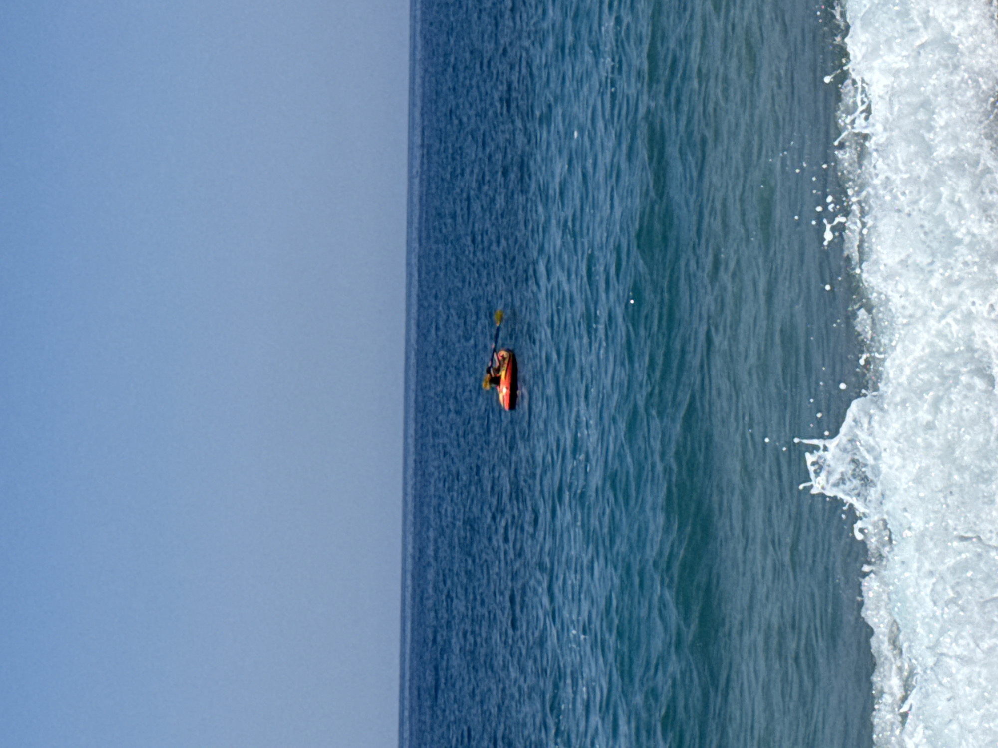 A bright orange inflatable raft with a person in it is seen in the middle of the azure blue water. The person is sitting in the raft and is holding a paddle, appearing to be kayaking. The raft is floating on the gentle waves. The scene is set against a clear blue sky and the deep blue expanse of the sea. The foreground features foamy white waves crashing onto the shore. The image appears to be taken on a sunny day in Alanya, Turkey, likely along its coastline.