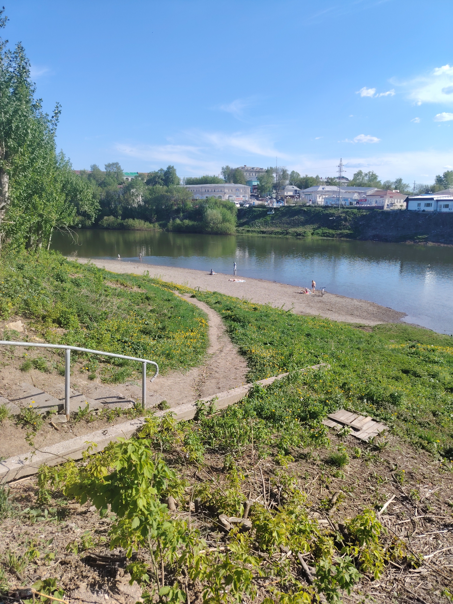 The image shows a landscape in Kungur, Russia, featuring a riverbank and surrounding terrain. In the foreground, there is a mix of vegetation, including grass, small shrubs, and some bare earth. A set of concrete steps with a metal railing leads down towards a sandy beach area along the river. Several people are on the beach, appearing to relax and enjoy the water. Across the river, there is a row of buildings, including some with white facades and others with more colorful roofs. The background reveals a blue sky with scattered clouds.