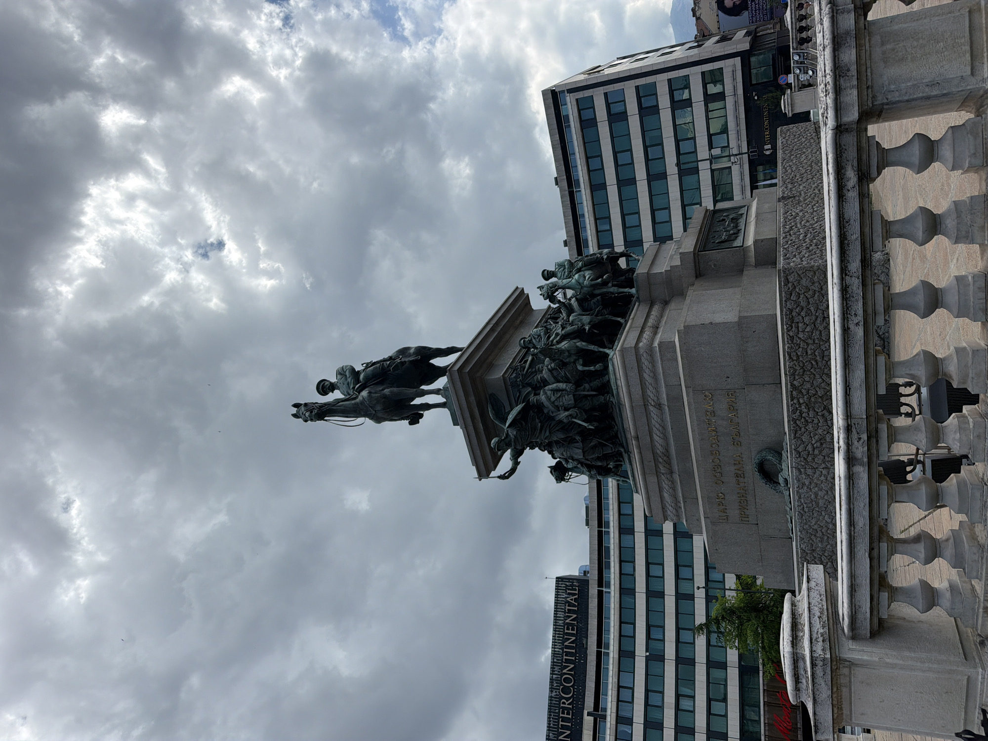 The image displays a prominent bronze equestrian monument under a heavily overcast, cloudy sky. The main statue depicts a rider on horseback, facing towards the right side of the frame, atop a grand stone pedestal. The base of the monument is intricately decorated with numerous smaller bronze figures, appearing to be soldiers or allegorical representations.
In the background, a modern, multi-story building with a facade dominated by windows is visible. Further to the right and slightly behind, another building features the distinct "INTERCONTINENTAL" hotel logo. To the far right, a decorative stone balustrade with classical spindles runs vertically.
Visible text on the monument's base, written in Cyrillic, reads: "ЦАРЬ ОСВОБОДИТЕЛЬ ПРИЗНАТЕЛНА БЪЛГАРИЯ," which translates to "Tsar Liberator Grateful Bulgaria." This text, along with the "Intercontinental" hotel sign, places the scene in Sofia, Bulgaria, identifying the monument as the Monument to the Tsar Liberator. The weather is cloudy and overcast, indicating daytime conditions without direct sunlight. No specific event or activity is taking place; the image is a static capture of the monument and its urban surroundings.