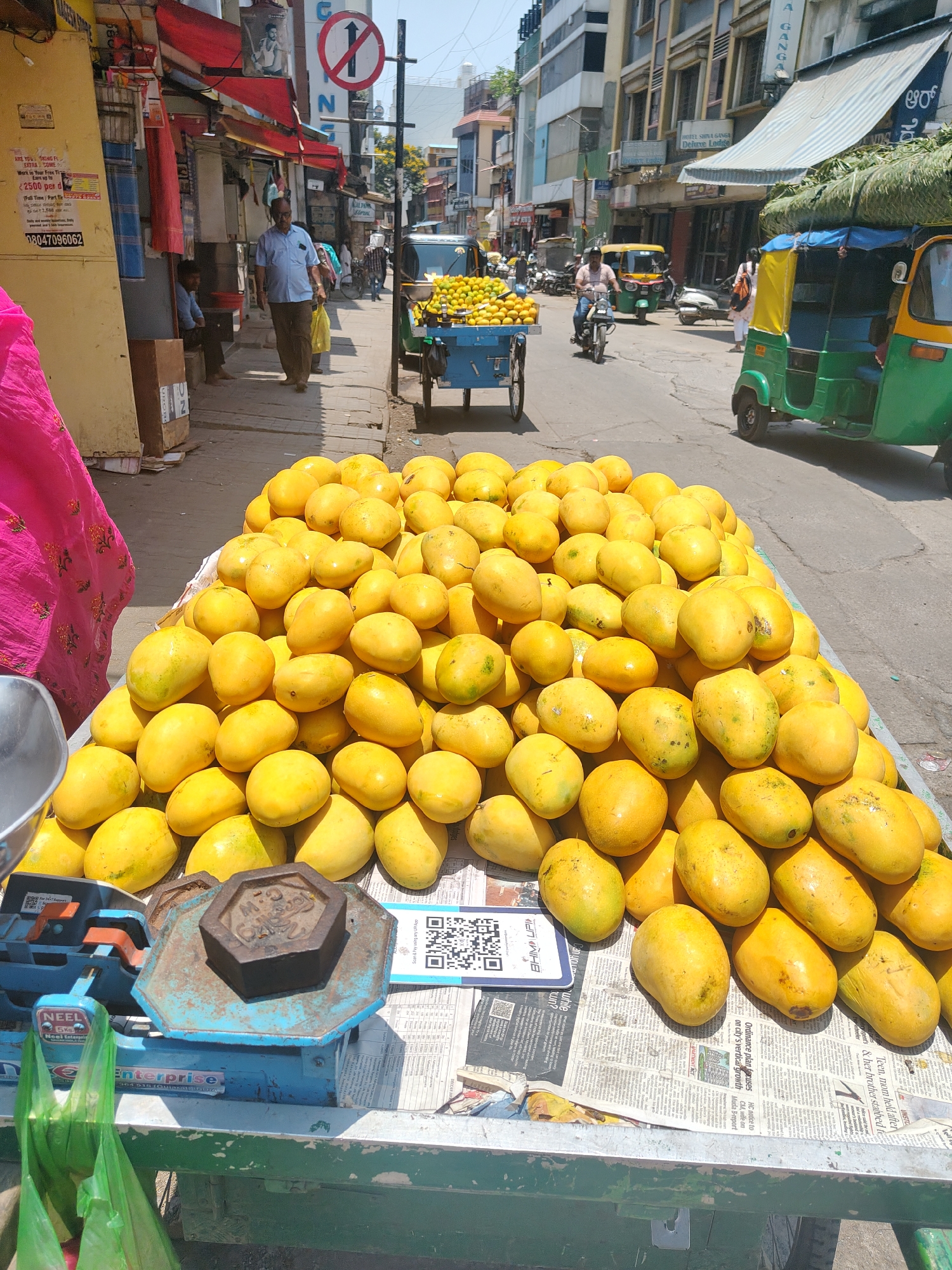 This media file depicts a vibrant street scene in Bengaluru, India, focusing on a roadside vendor's stall laden with mangoes. A large pile of bright yellow, ripe mangoes occupies the foreground, displayed on a green and blue cart. Adjacent to the mangoes, a traditional blue mechanical weighing scale, branded "NEEL Enterprise," is visible with a dark, hexagonal weight. Green plastic bags are placed nearby, ready for use. Newspapers, serving as a base for the display, feature visible headlines including "APARTMENT ACT Ordinance plan focuses on city's vertical growth" and "Teen, mom held after gent's & her brother stabbed fo." A QR code is present on the newspaper, alongside the prompt "Scan & Pay using any UPI App," indicating digital payment options.
The image captures a sunny, clear daytime, evident from the strong shadows. In the background, a busy street unfolds with several people and vehicles. A man in a light blue shirt walks on the sidewalk past shops with red awnings. Farther down the street, another vendor's cart, piled with oranges, is visible. Auto-rickshaws are a prominent feature, with a green and yellow one on the right, and a yellow one in the distance alongside a person on a motorcycle. Multi-story buildings line the street, displaying signs for businesses such as "HOTEL SHIVA GANGA Deluxe Lodge" and "AGANGA," with Kannada script visible on some signage. A red circular "No Entry" or similar traffic sign is also visible on a pole. The scene portrays a lively urban market environment, characteristic of an Indian city, where traditional vending meets modern commerce.