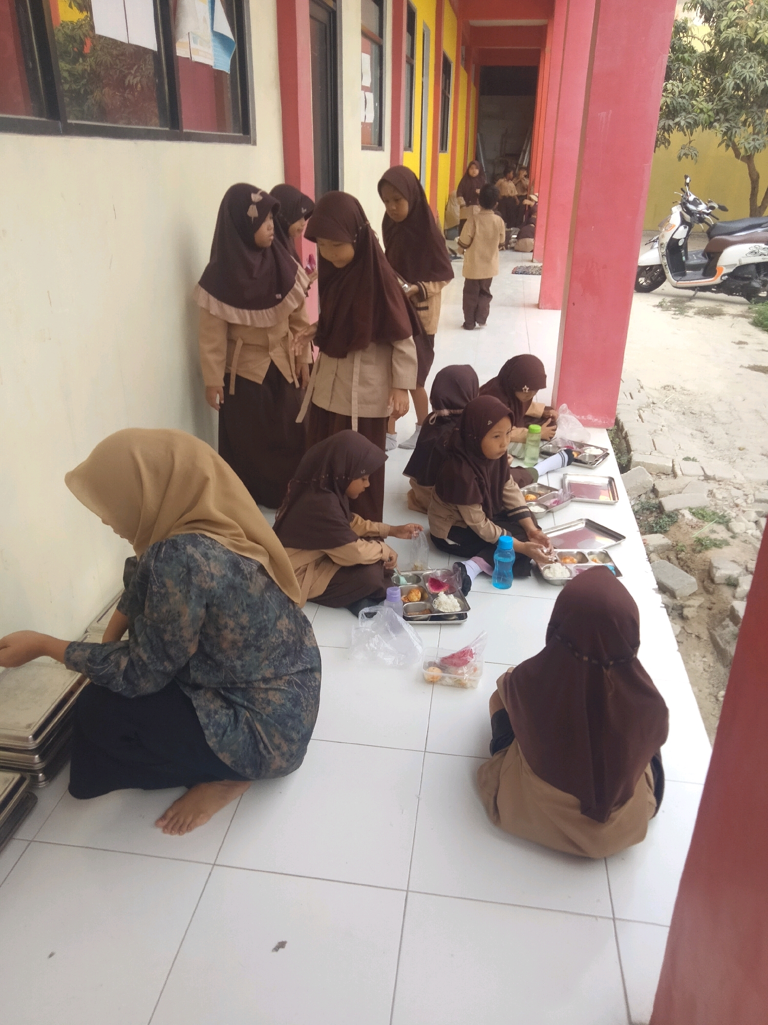 In the city of Suka Sari, Indonesia, a group of female students wearing brown hijabs and school uniforms are seated on a tiled floor outdoors, presumably during a break or mealtime. The students are arranged in two main groups. One group of four students is seated on the right side of the image, facing towards the left, engaged with trays of food and drinks. Another group of two students is situated in the foreground on the left, also appearing to be interacting with food. A scooter is parked in the background to the right of the students. The environment appears to be a school corridor or walkway, with a pink-colored wall on the right and a building with windows on the left. The ground is covered in white tiles.